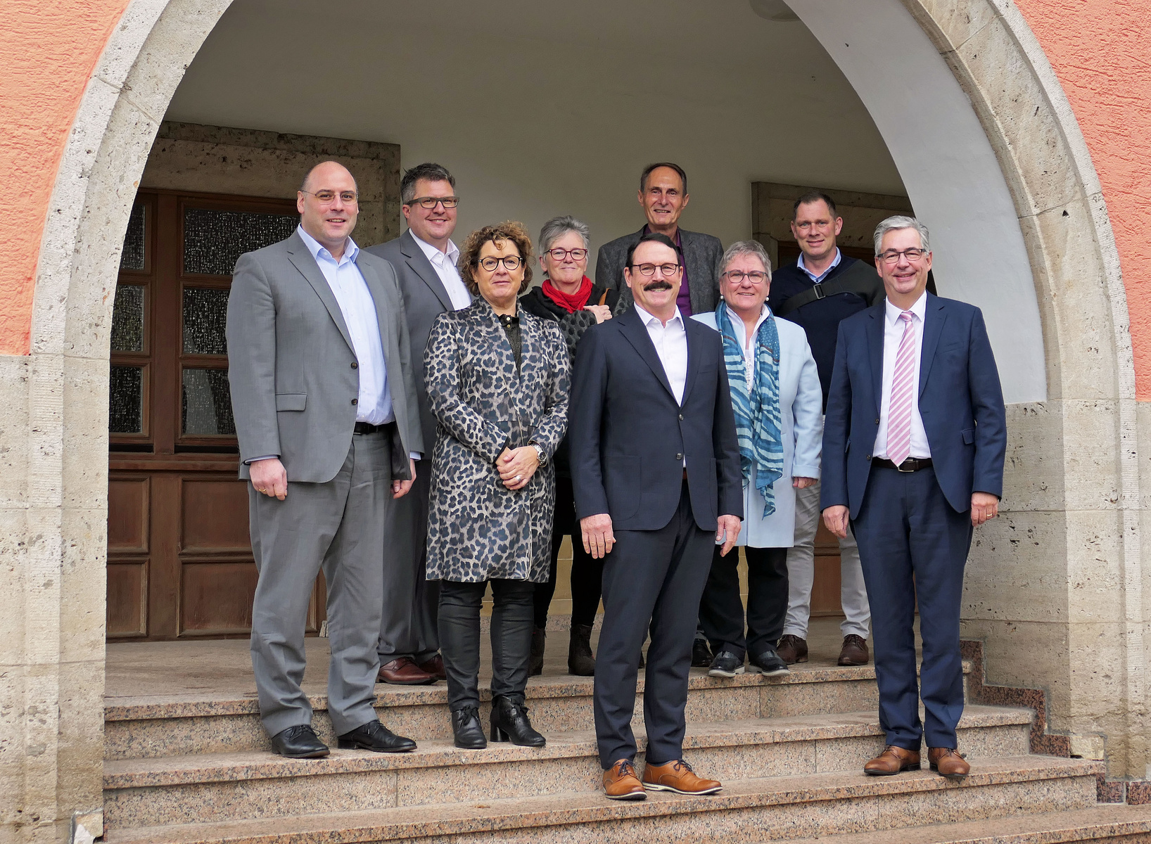 Un groupe de neuf personnes se tient en posant sur des marches devant une entrée d'immeuble, souriantes. Elles sont vêtues de manière professionnelle, certaines avec des vêtements à motifs. L'ambiance semble conviviale et chaleureuse, suggérant une réunion ou un événement formel.