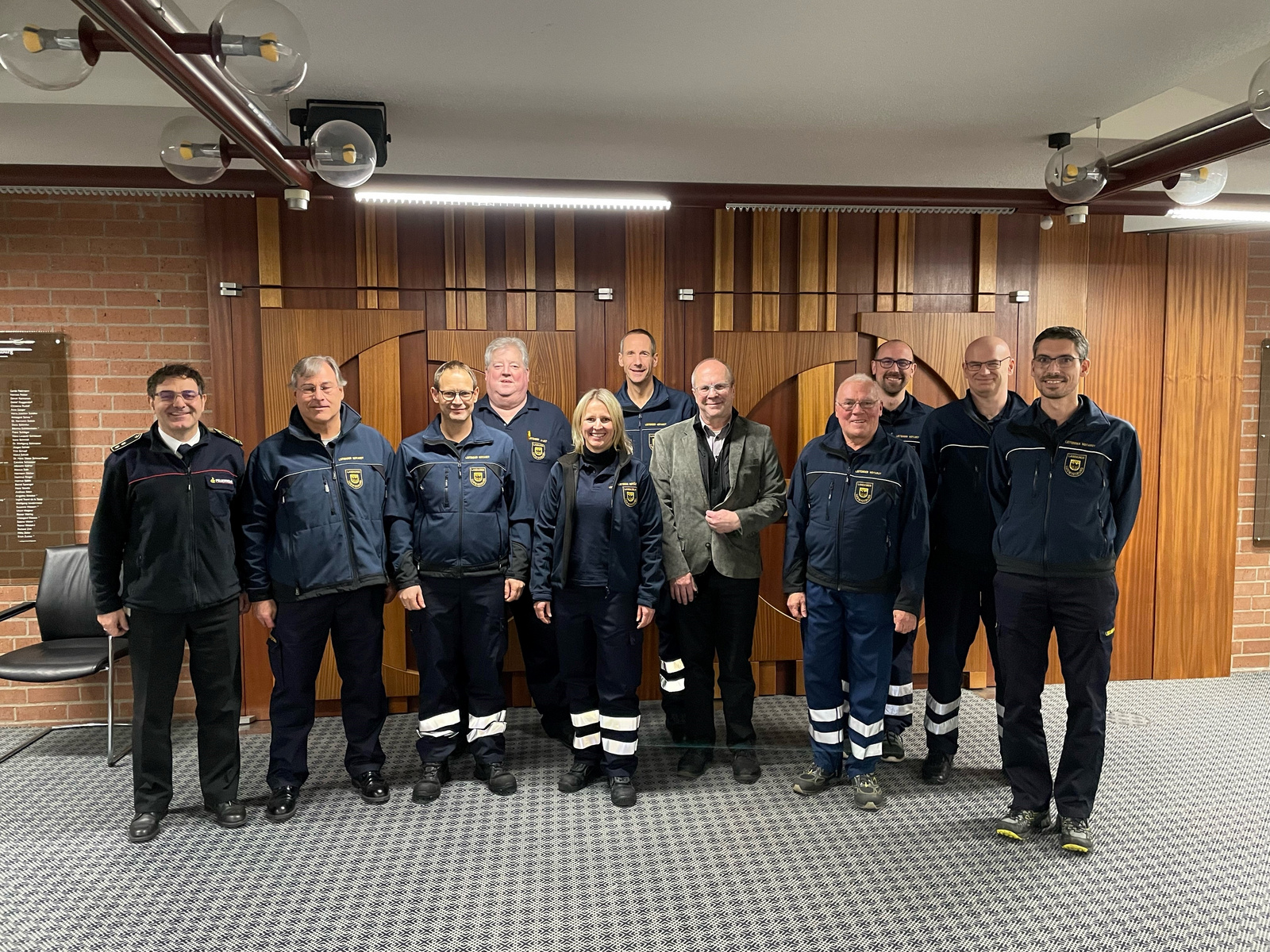 Un groupe de douze personnes, dont des membres en uniforme, pose ensemble. Ils se tiennent côte à côte avec des sourires, sur un fond en bois. Certains portent des écussons ou logos, indiquant leur affiliation professionnelle. L'image illustre la camaraderie et le travail d'équipe.