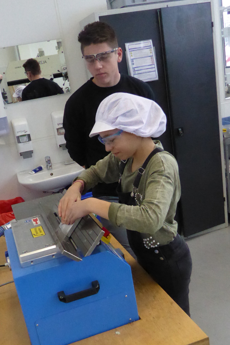 Eine Person, die eine Schutzbrille trägt, beobachtet eine zweite Person, die auf einem Tisch an einer Maschine arbeitet. Die Maschine ist blau und wird zur Bearbeitung eines Werkstücks verwendet. Beide Personen tragen geeignete Schutzkleidung.