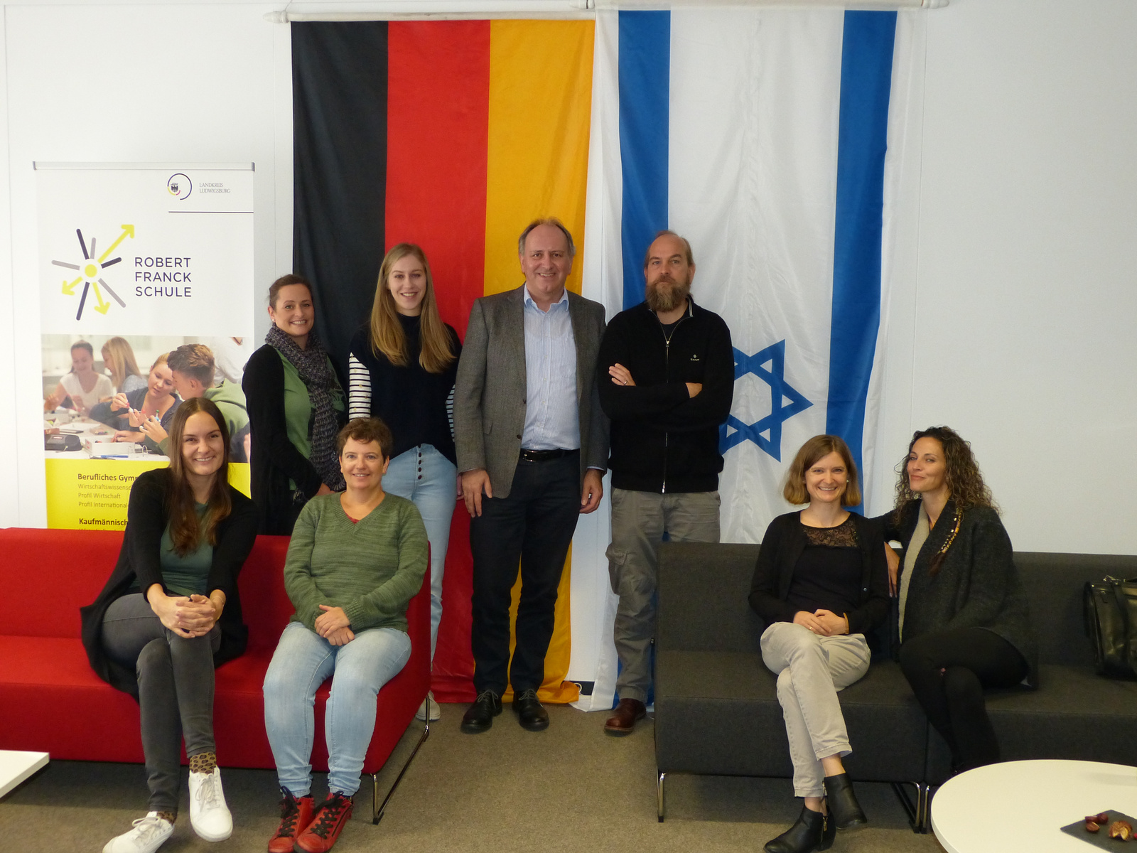 Grupp portrait in einem Schulungsraum, in dem sieben Personen stehen und sitzen. Im Hintergrund sind die Flaggen Deutschlands und Israels zu sehen, sowie ein Schild der Robert Franck Schule. Alle Personen lächeln und scheinen an einem gemeinsamen Projekt beteiligt zu sein.