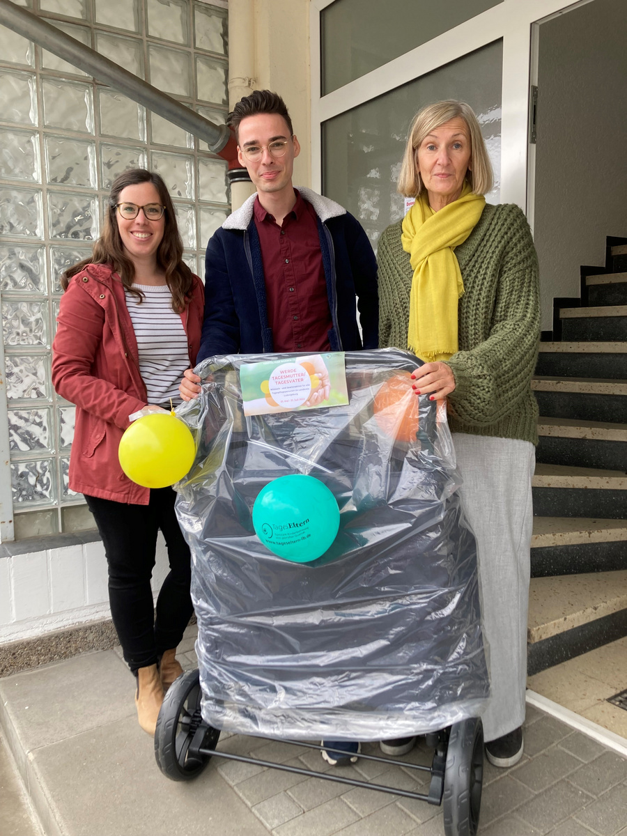 Drei Personen stehen in einem Hausflur vor einem Kinderwagen, der mit Luftballons und einigem Geschenkpapier dekoriert ist. Die Frauen tragen unterschiedliche Oberbekleidung. Der Mann in der Mitte lächelt und hält den Kinderwagen fest. Es scheint eine Geschenkübergabe oder Feierlichkeit zu sein.