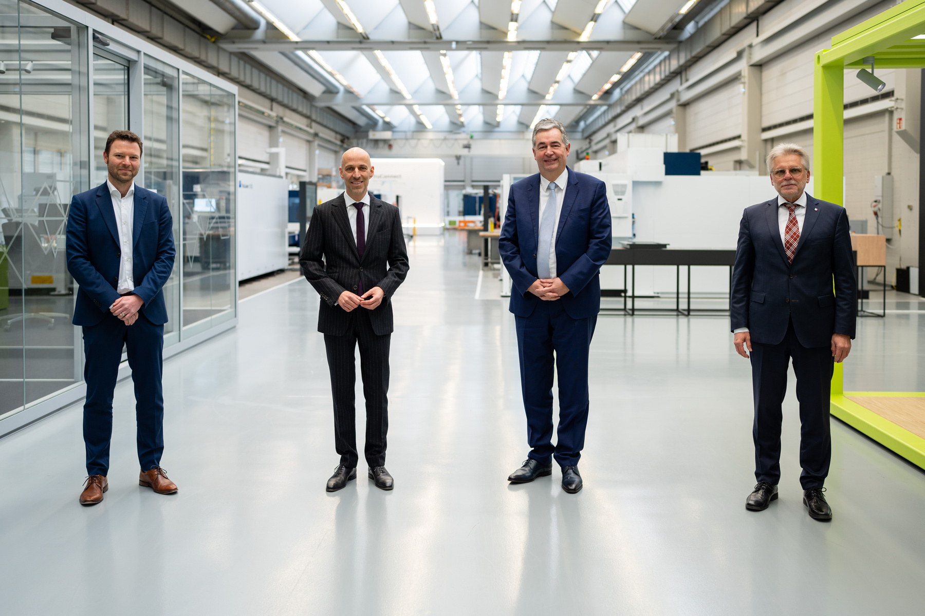 Four men in business attire stand in a spacious, well-lit industrial setting. They appear to be engaged in a formal event, possibly a meeting or announcement, with machinery visible in the background. Each person has a confident posture and is looking towards the camera.