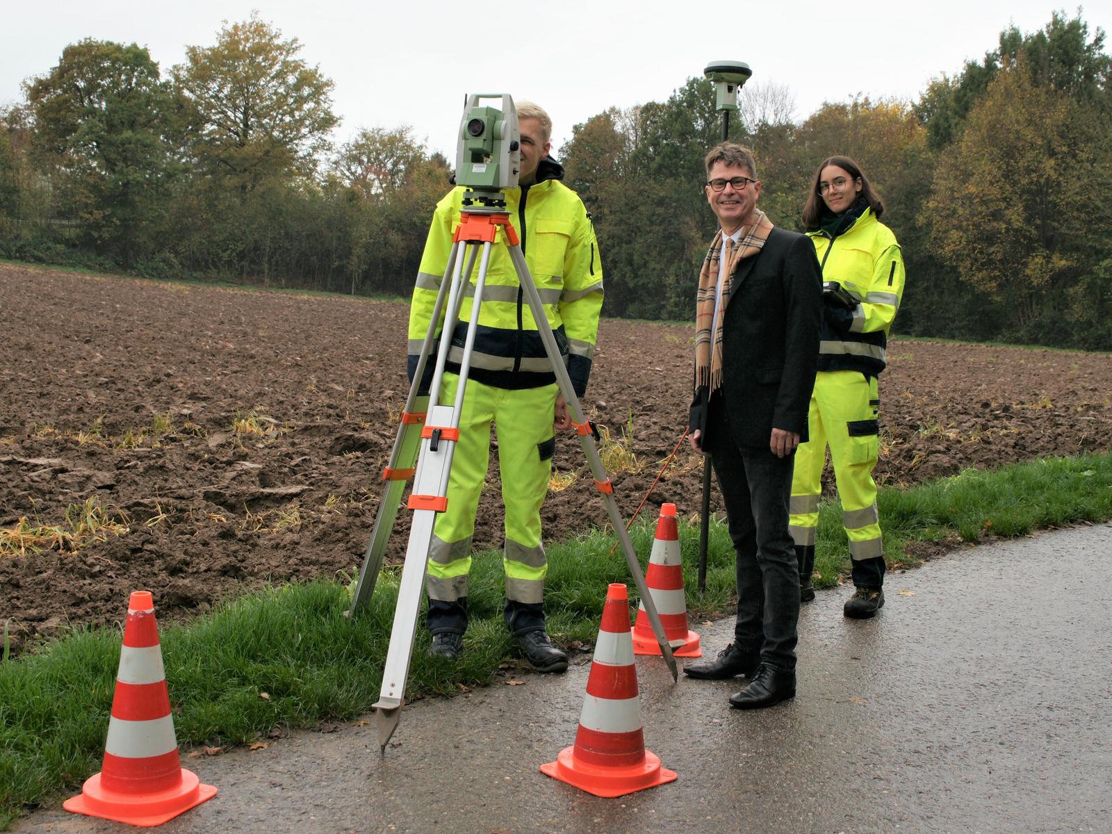 Drei Personen stehen auf einem Feldweg. Zwei in auffälligen, gelben Arbeitsanzügen halten ein Vermessungsgerät, während der dritte Mann in einem dunklen Anzug daneben steht. Orangefarbene Verkehrshütchen sind im Vordergrund sichtbar. Bäume im Hintergrund zeigen die Herbstsaison.