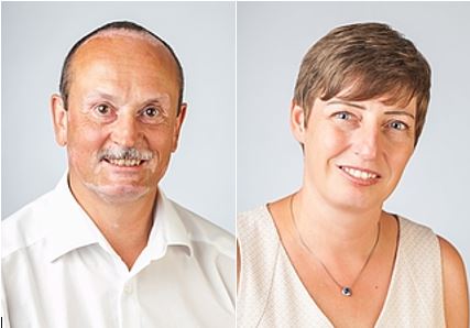 Das Bild zeigt zwei Personen: einen Mann mit Schnurrbart und einem weißen Hemd links, und eine Frau mit kurzem, braunem Haar und einem hellen Oberteil rechts. Beide lächeln freundlich in die Kamera vor einem neutralen Hintergrund.