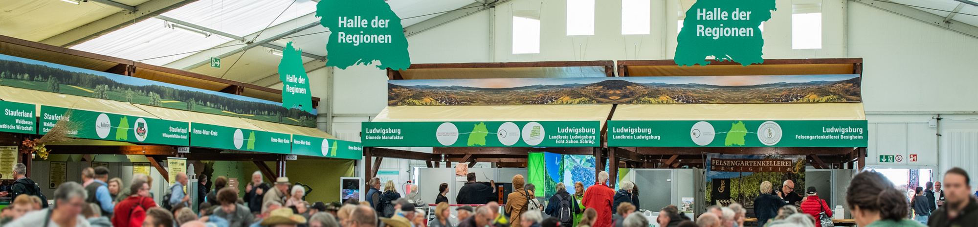 Ein überfüllter Veranstaltungsraum mit Ständen und Anzeigetafeln, die die "Halle der Regionen" markieren. Die Menschen stehen zusammen, während einige an den Ständen einkaufen oder informieren. Helle Beleuchtung und ein großes Zeltdach sind sichtbar. (Text KI generiert)