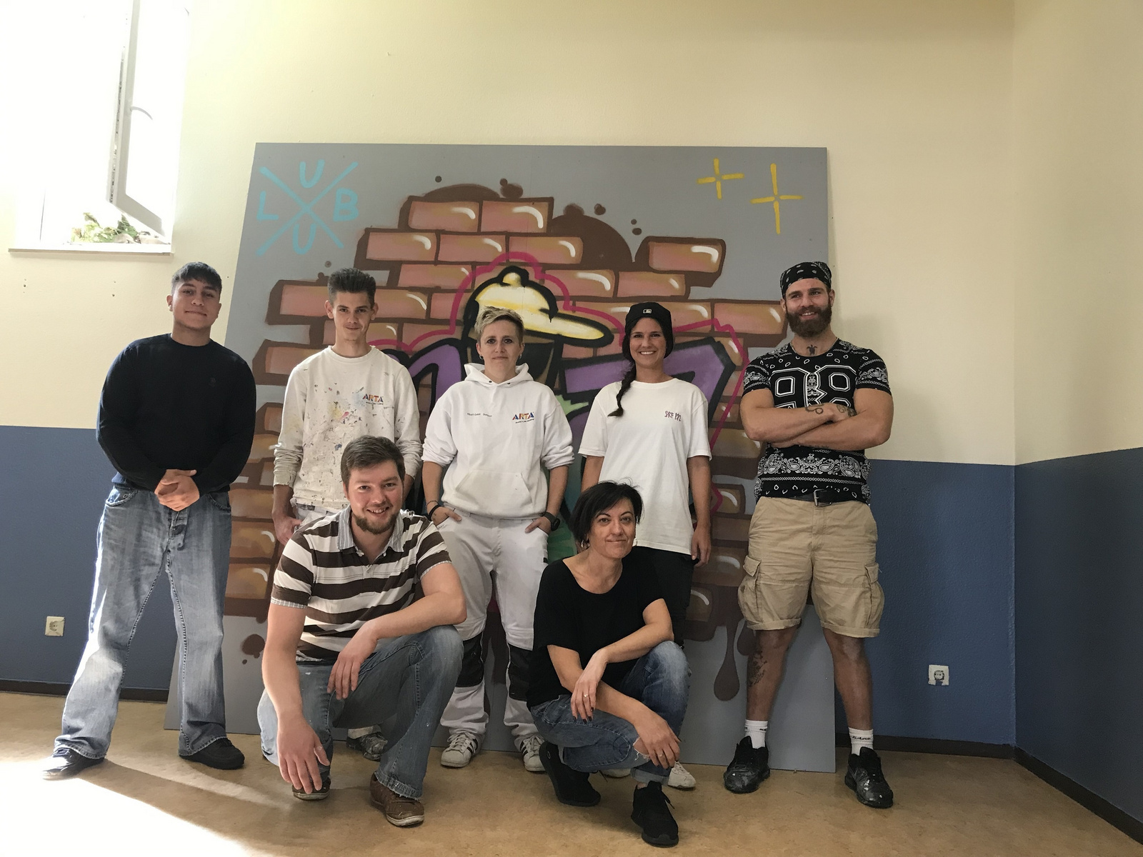 Eine Gruppe von sieben Personen steht vor einem Wandgemälde mit einer Mauer aus Ziegelsteinen und bunten grafischen Elementen. Die Teilnehmer sind unterschiedlich gekleidet und lächeln in die Kamera, während sie in zwei Reihen positioniert sind.