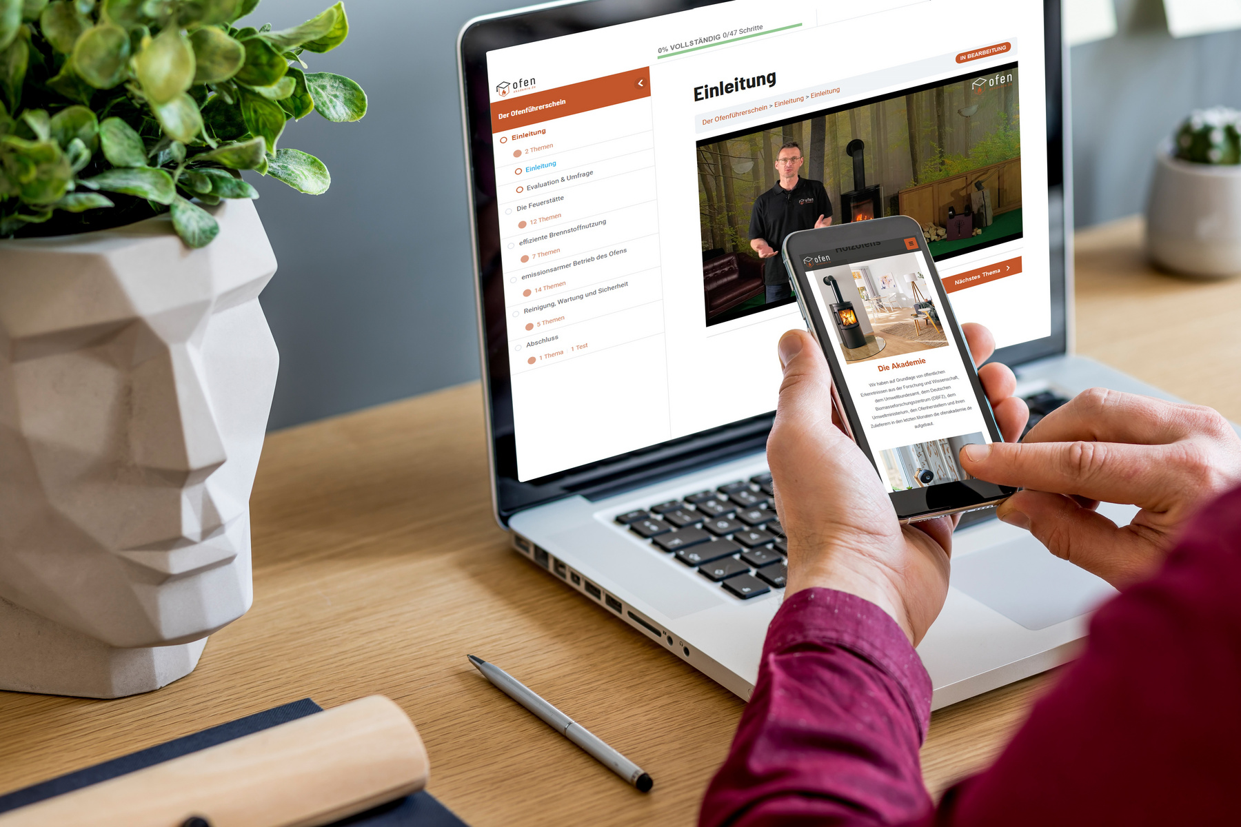 Ein Mensch hält ein Smartphone in der Hand, während er auf einen Laptop schaut. Auf dem Laptop-Bildschirm ist eine Webseite mit dem Titel „Einleitung“ sichtbar. Im Hintergrund steht eine Pflanze in einem modernen Topf. Der Arbeitsplatz ist ordentlich und minimalistisch eingerichtet.