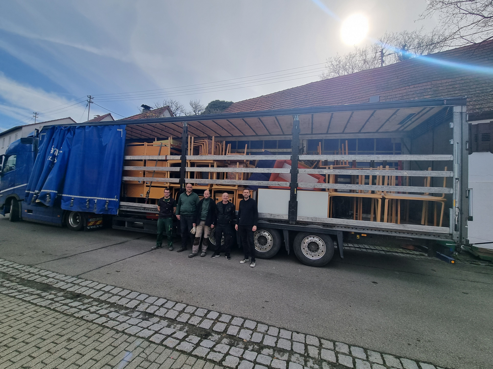 Ein Lkw steht auf einer Straße, beladen mit Tischen und Stühlen. Vor dem Lkw stehen fünf Personen in Arbeitskleidung. Im Hintergrund sind ein paar Gebäude und ein blauer Himmel zu sehen.