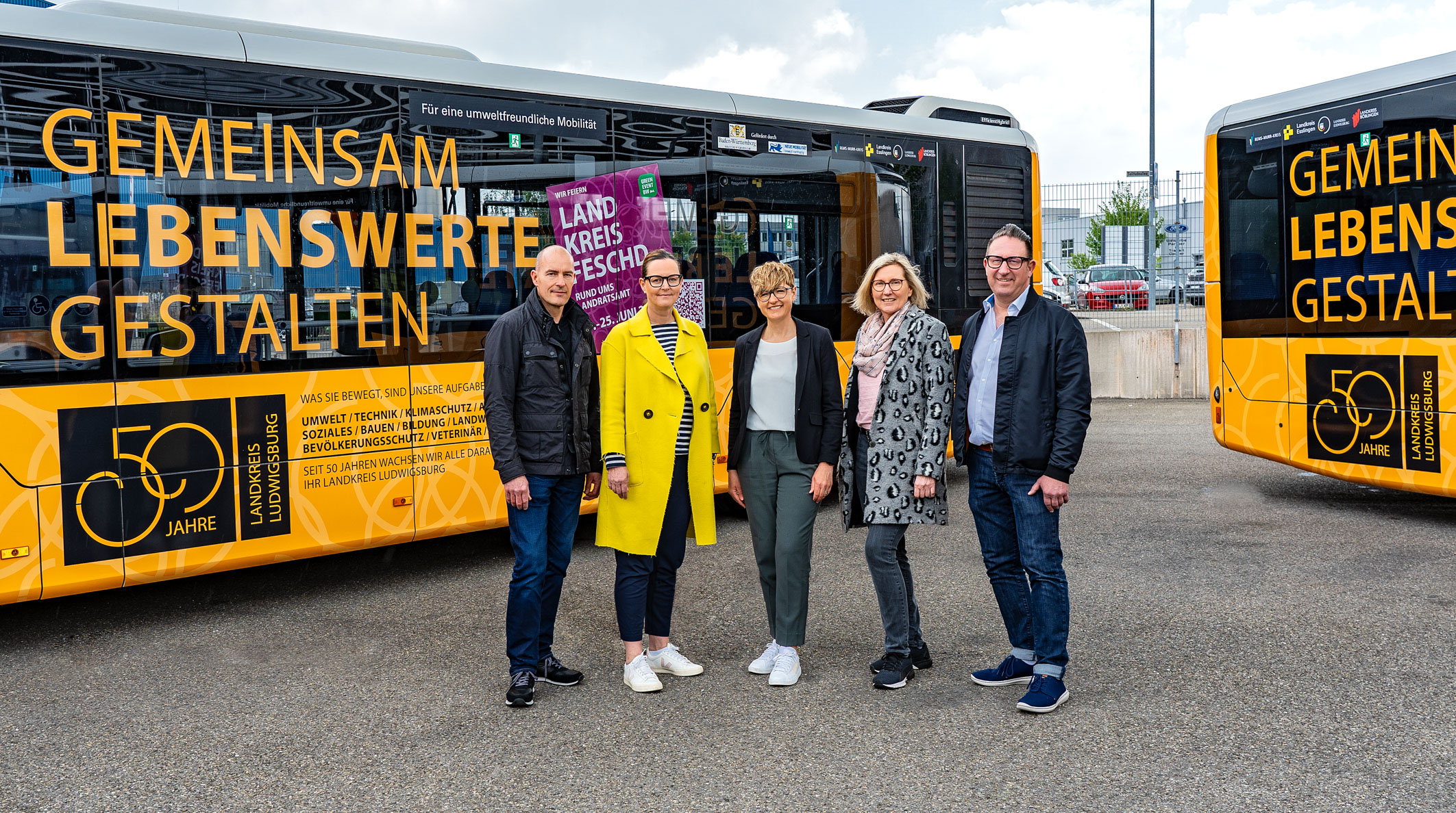 Fünf Personen stehen vor zwei Bussen mit einer Werbebotschaft. Die Botschaft auf den Bussen lautet „Gemeinsam lebenswerte gestalten“. Im Hintergrund sind Wolken und der Parkplatz zu sehen. Die Szene vermittelt ein Gefühl von Zusammenarbeit und Gemeinschaftsprojekten.