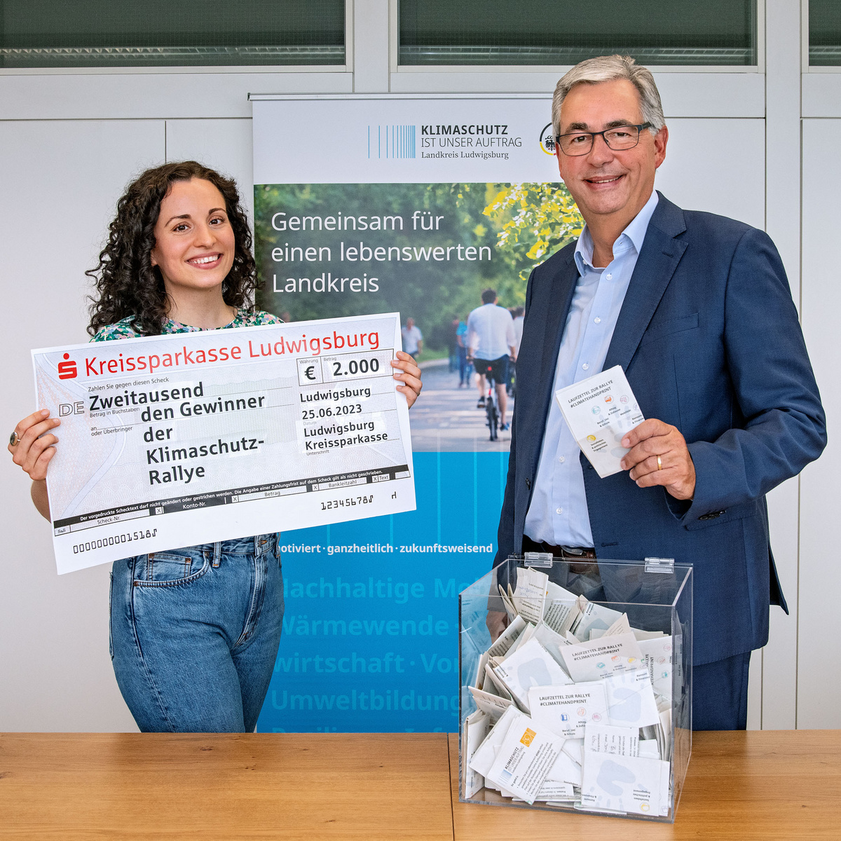Due persone sorridenti, una donna e un uomo, sono in piedi davanti a un poster. La donna tiene un grande assegno che indica un premio di 2.000 euro per un concorso di tutela climatica, mentre l'uomo mostra un modulo. Sullo sfondo, informazioni sul "Klimaschutz Rallye".