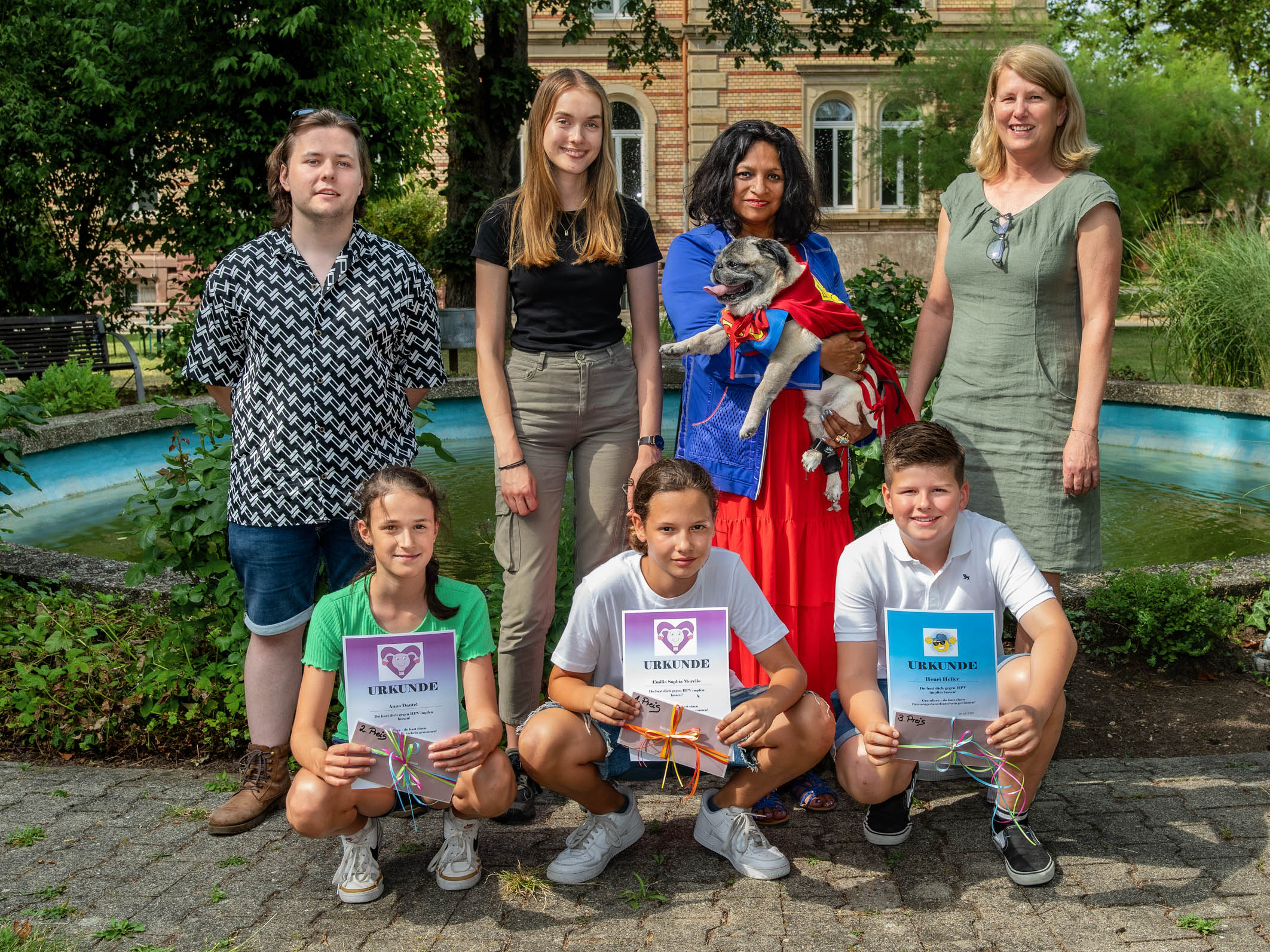 Eine Gruppe von sieben Personen steht um einen Brunnen im Freien. Die Erwachsenen stehen hinter den Kindern, die Urkunden mit dem Schriftzug "Danke" in der Hand halten. Eine Person hält einen kleinen Hund, während die anderen lächeln und in einer freundlichen Atmosphäre posieren.