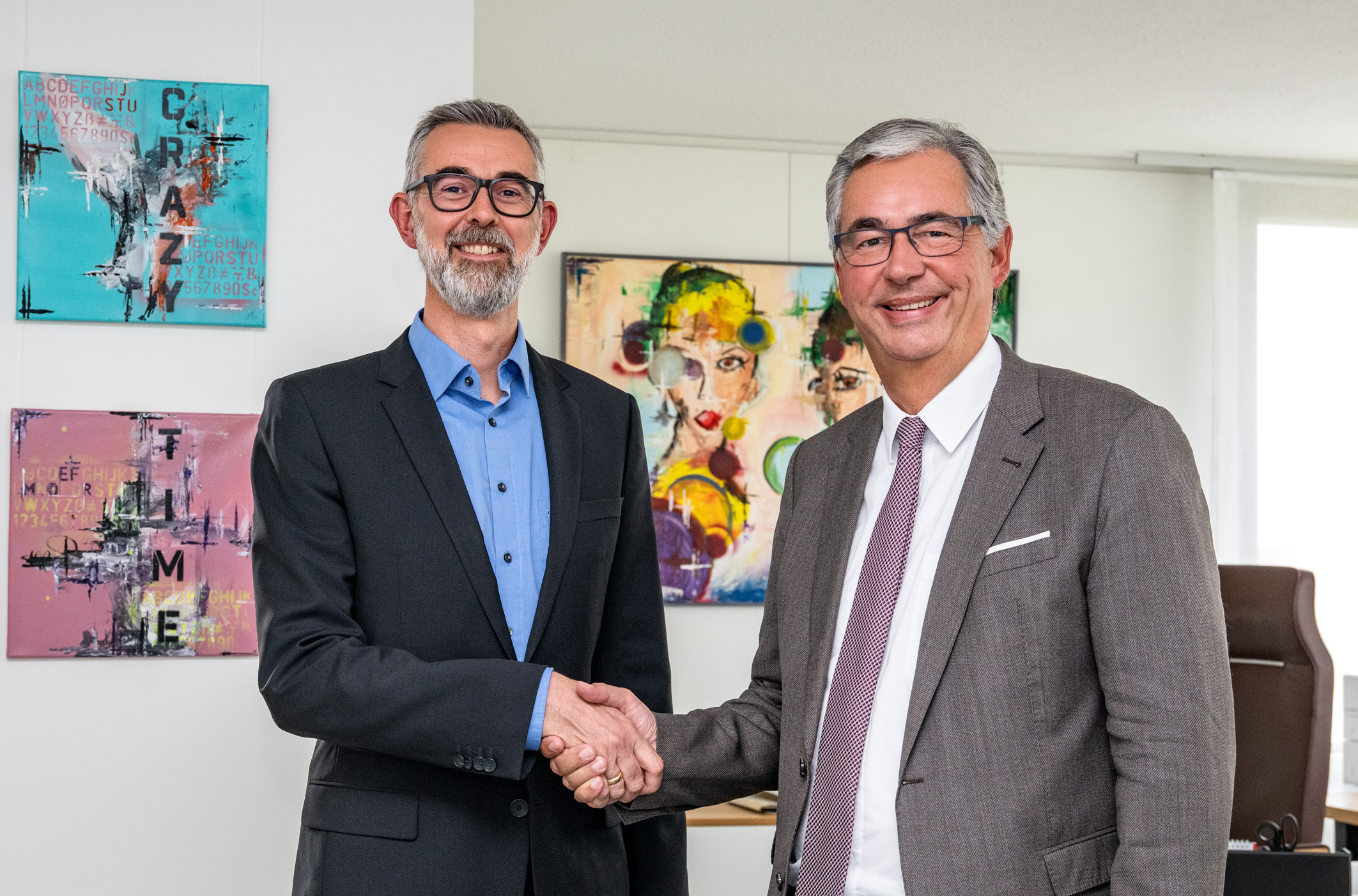 Zwei Männer in Anzügen schütteln sich die Hände in einem Büro. Im Hintergrund sind bunte Kunstwerke an der Wand zu sehen. Die Szene vermittelt einen formellen, geschäftlichen Austausch oder eine Vereinbarung.