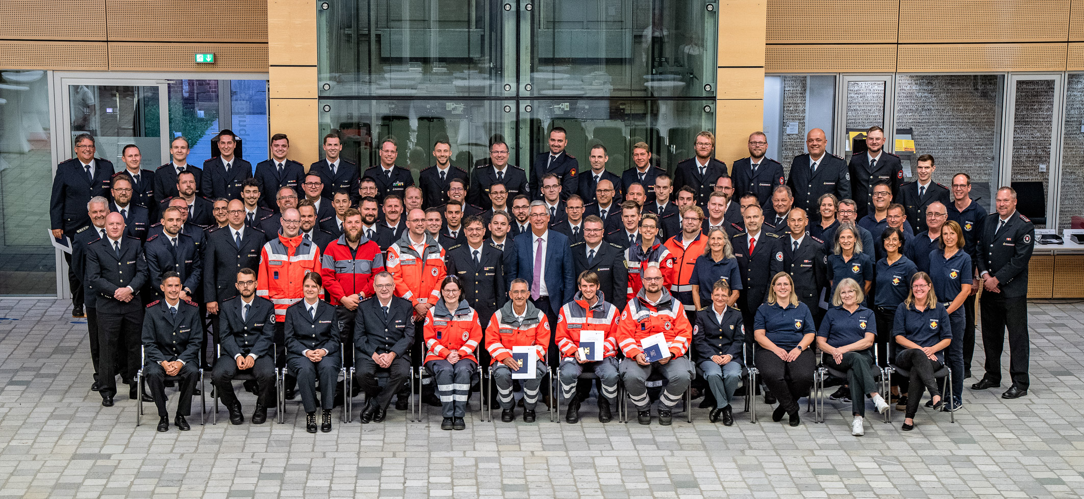 Gruppenfoto von etwa 75 Personen in verschiedenen Uniformen, darunter Feuerwehr, Rettungsdienst und Zivilkleidung. Sie stehen vor einem modernen Gebäude. Die Atmosphäre ist freundlich und professionell, mit dem Fokus auf Teamarbeit und Gemeinschaft.