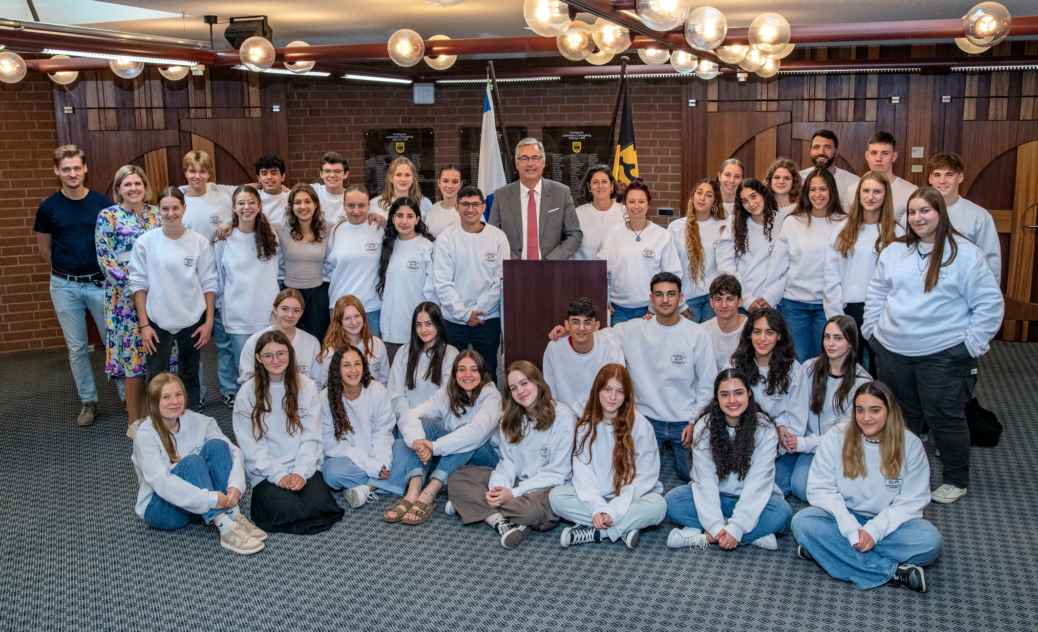 Eine Gruppe von etwa 40 Jugendlichen in weißen Sweatshirts steht vor einem Rednerpult. Dahinter befindet sich ein Mann in Anzug und Krawatte, der spricht. Im Hintergrund sind Fahnen und eine Wand mit Holzstruktur sichtbar. Die Atmosphäre wirkt freundlich und gemeinschaftlich.