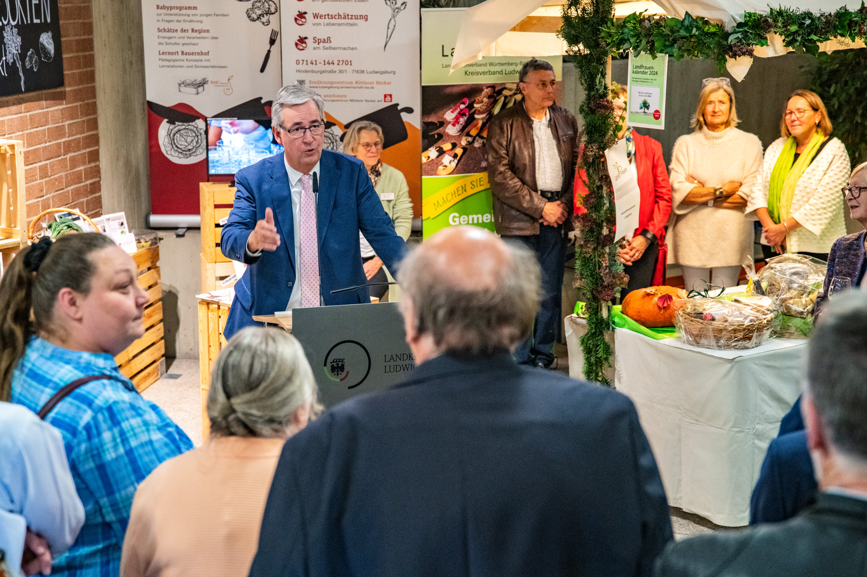 Ein Mann in Anzug spricht an einem Podium vor einer Gruppe von Menschen. Im Hintergrund sind Stände mit Informationen und Produkten zu sehen. Die Anwesenden hören aufmerksam zu, während er seine Botschaft vermittelt.