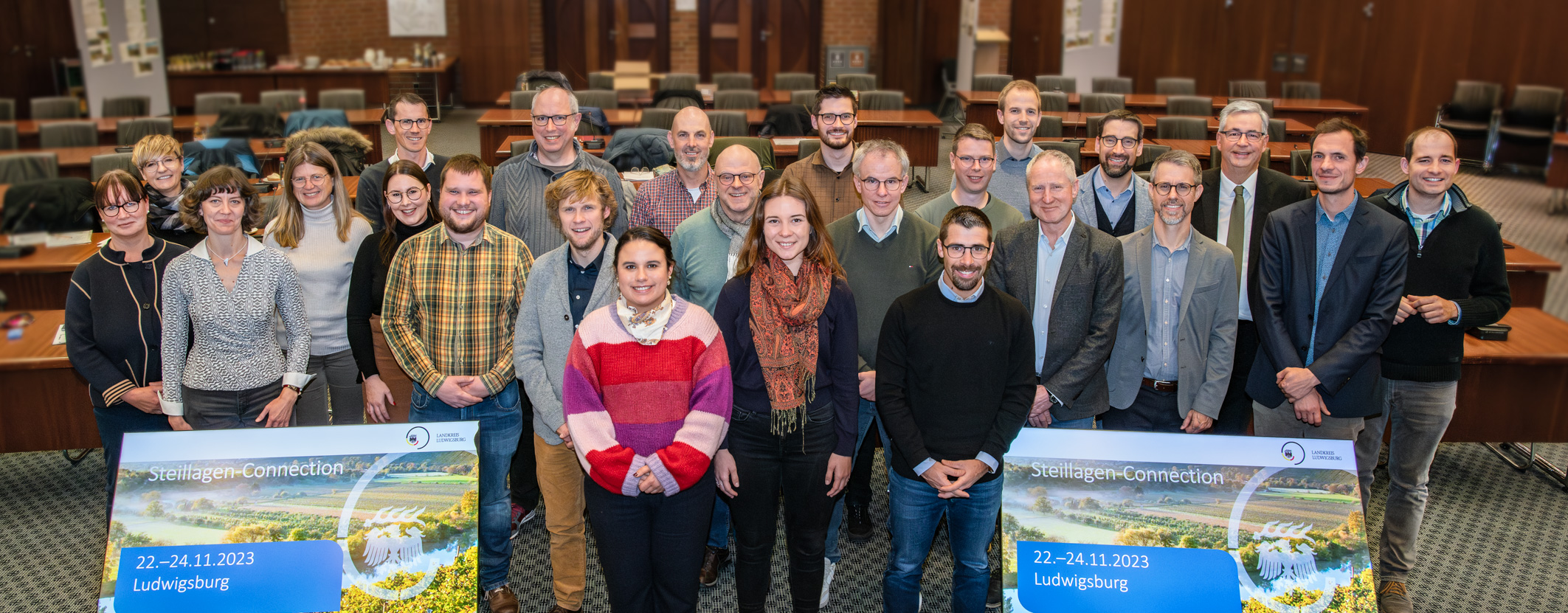 Eine Gruppe von Menschen steht in einem großen Raum. Sie sind in einem semi-formellen Umfang gekleidet und lächeln in die Kamera. Im Vordergrund sind zwei große Banner mit dem Titel „Connecting Connections“ und dem Datum 22.-24.11.2023 in Ludwigsburg zu sehen.