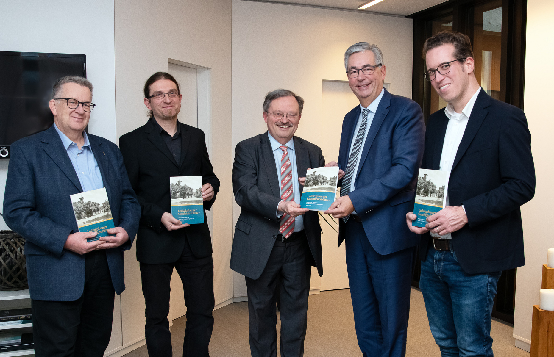 Vier Männer stehen in einem Raum und halten jeweils ein Buch in der Hand. Sie scheinen sich über die Bücher auszutauschen, während ein Mann in der Mitte ein Buch an einen anderen Mann übergibt. Alle wirken lächelnd und freundlich.