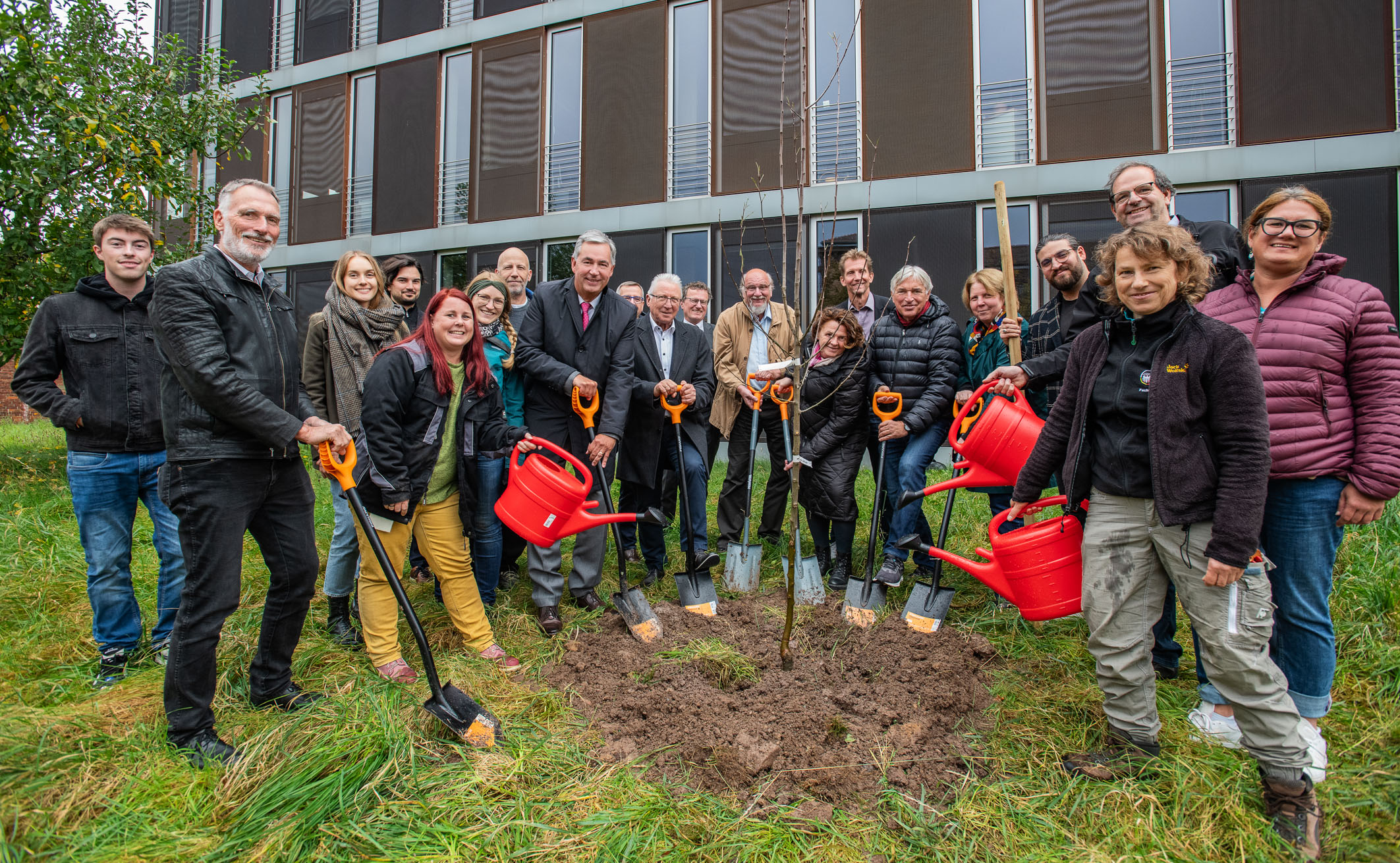 Eine Gruppe von 15 Personen steht um eine frisch gegrabene Erdgrube, bereit, Bäume zu pflanzen. Einige halten Schaufeln, andere Gießkanne. Im Hintergrund ist ein modernes Gebäude zu sehen. Alle wirken konzentriert und engagiert bei der Pflanzaktion.