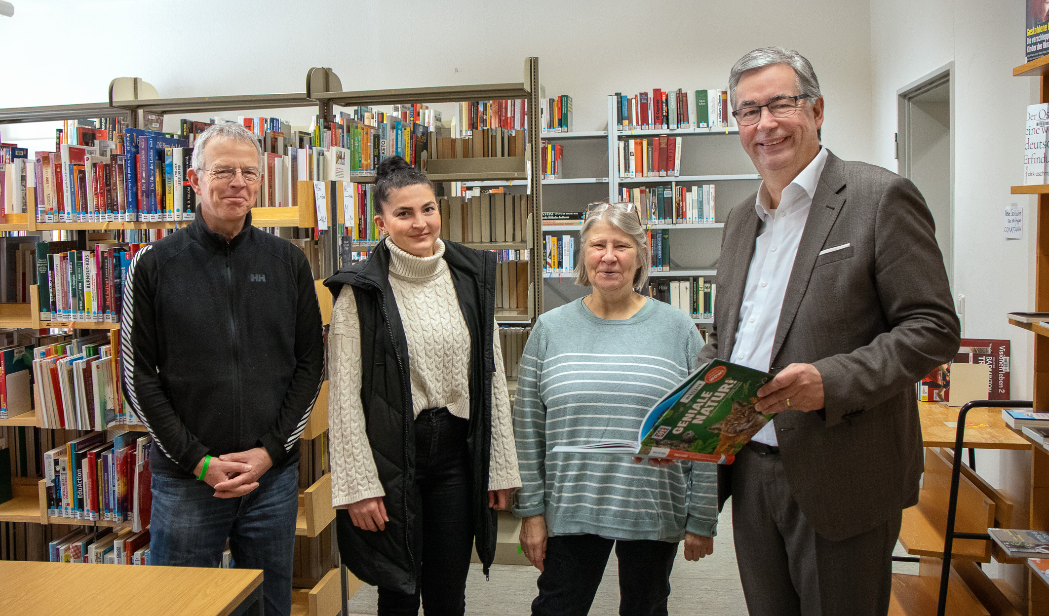 Vier Personen stehen in einer Bibliothek zwischen Bücherregalen. Der Mann rechts hält ein Buch in der Hand. Die anderen Personen lächeln und stehen nebeneinander, während sie freundlich in die Kamera blicken. Die Atmosphäre wirkt einladend und gemeinschaftlich.