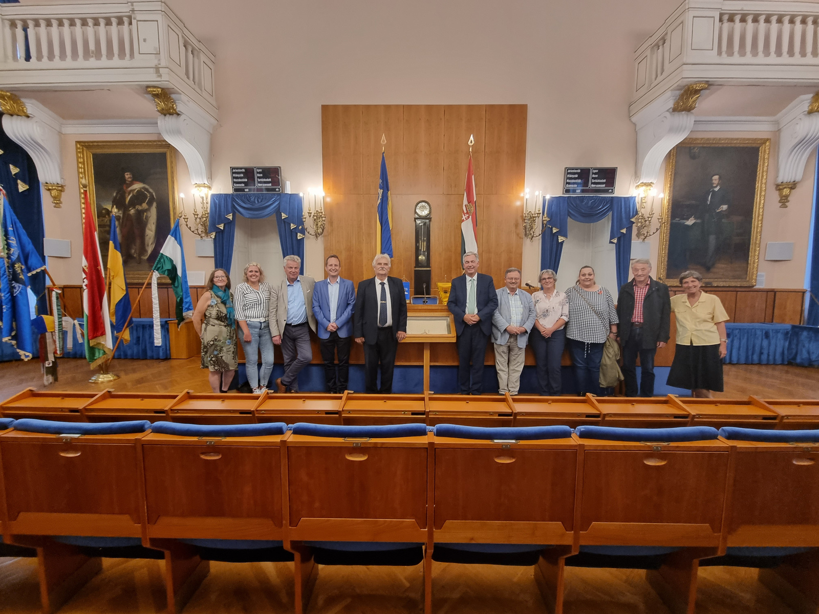Eine Gruppe von zwölf Personen steht in einem Saal mit Holzmöbeln und Flaggen. Im Hintergrund sind große Gemälde an den Wänden sichtbar. Die Personen lächeln und posieren zusammen, während sie sich für ein Foto aufstellen.