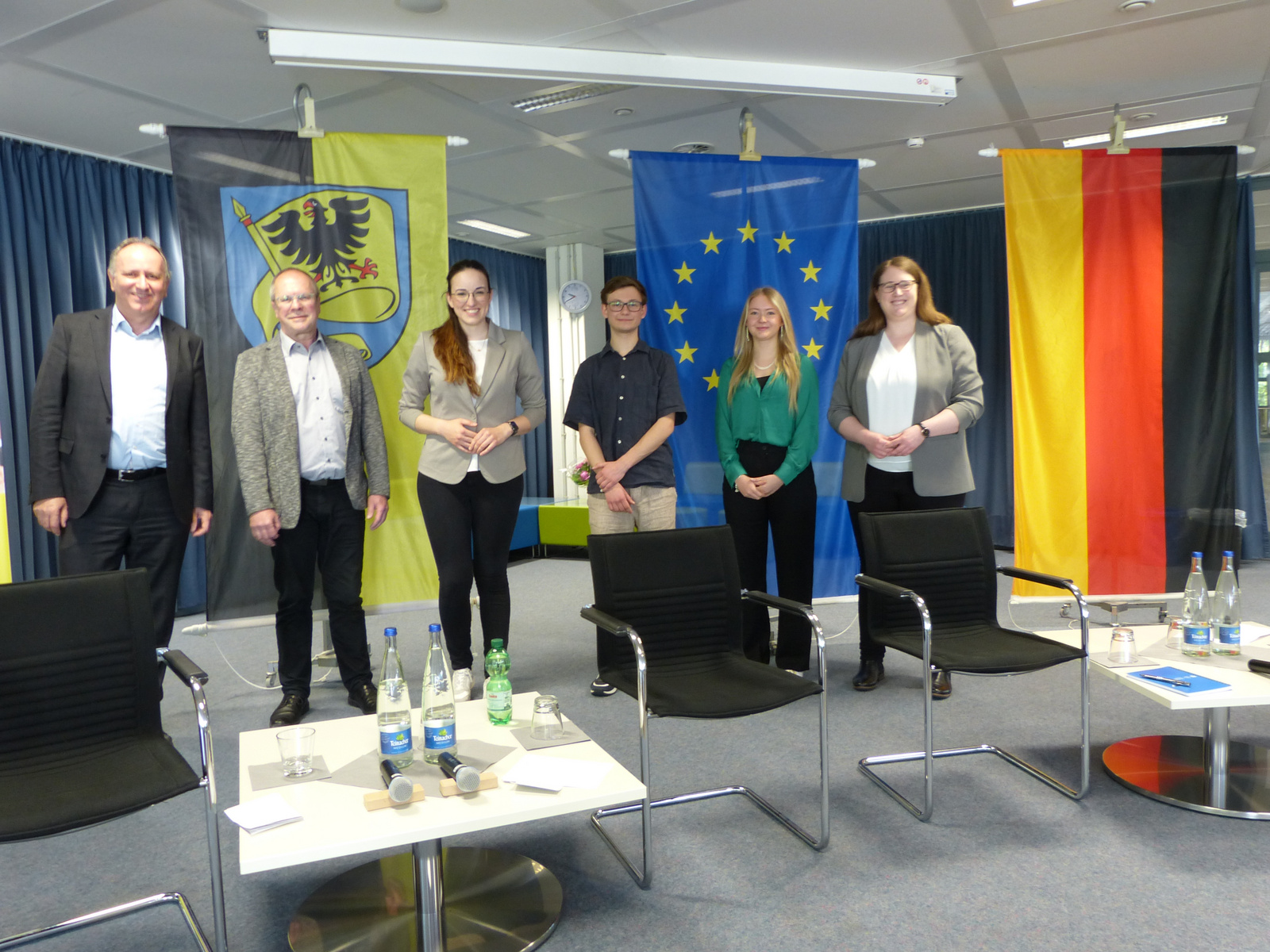 Sechs Personen stehen in einem Raum mit Fahnen im Hintergrund, darunter die deutsche und die europäische Flagge. Sie scheinen an einer Veranstaltung teilzunehmen. Auf einem Tisch stehen Wasserflaschen und Mikrofone.