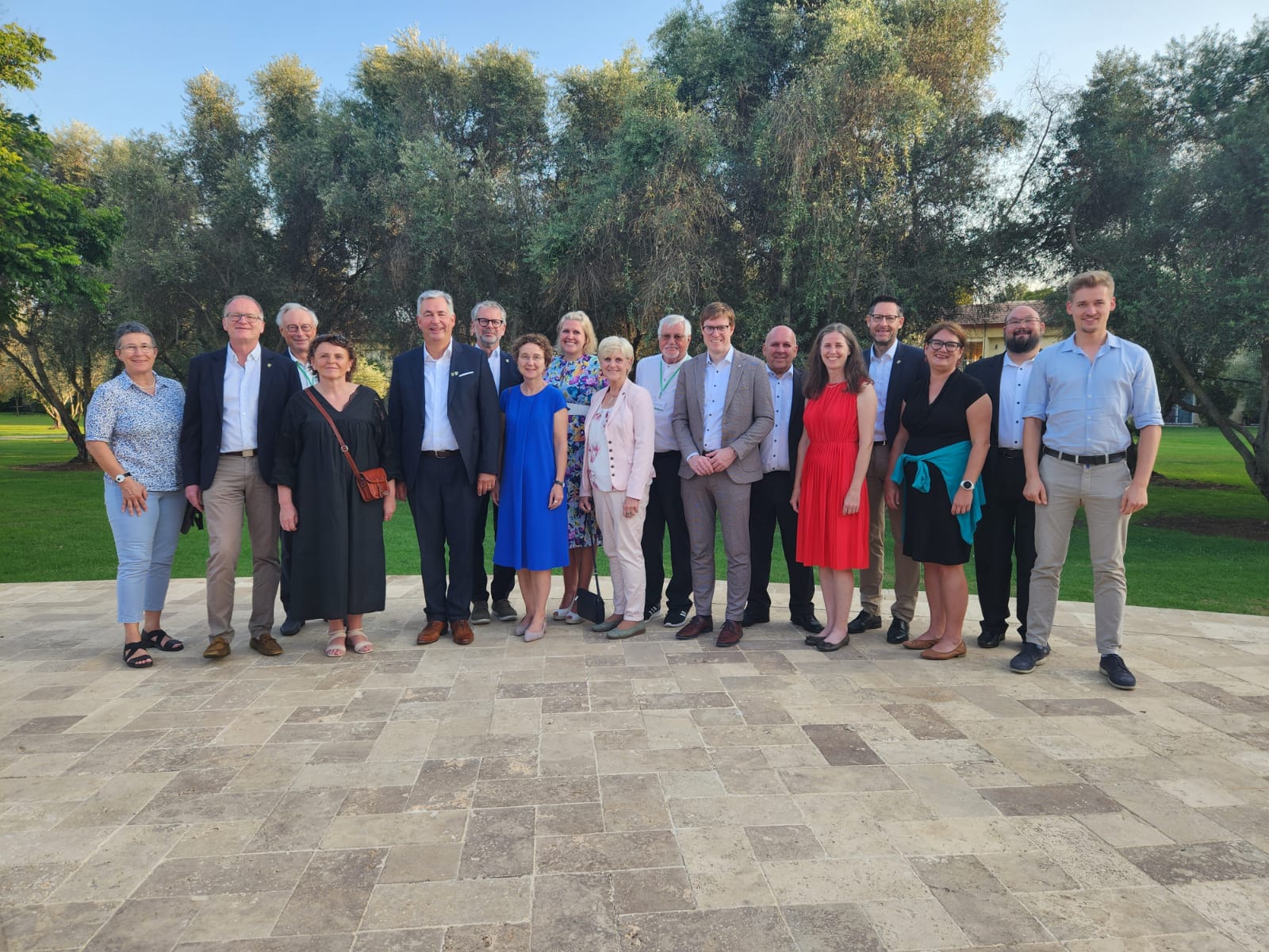 Eine Gruppe von 18 Personen steht auf einem eleganten Steinplatz im Freien, umgeben von Bäumen. Die Anwesenden tragen unterschiedliche, festliche Kleidung. Sie lächeln und posieren für ein Gruppenfoto, das eine fröhliche Atmosphäre vermittelt.
