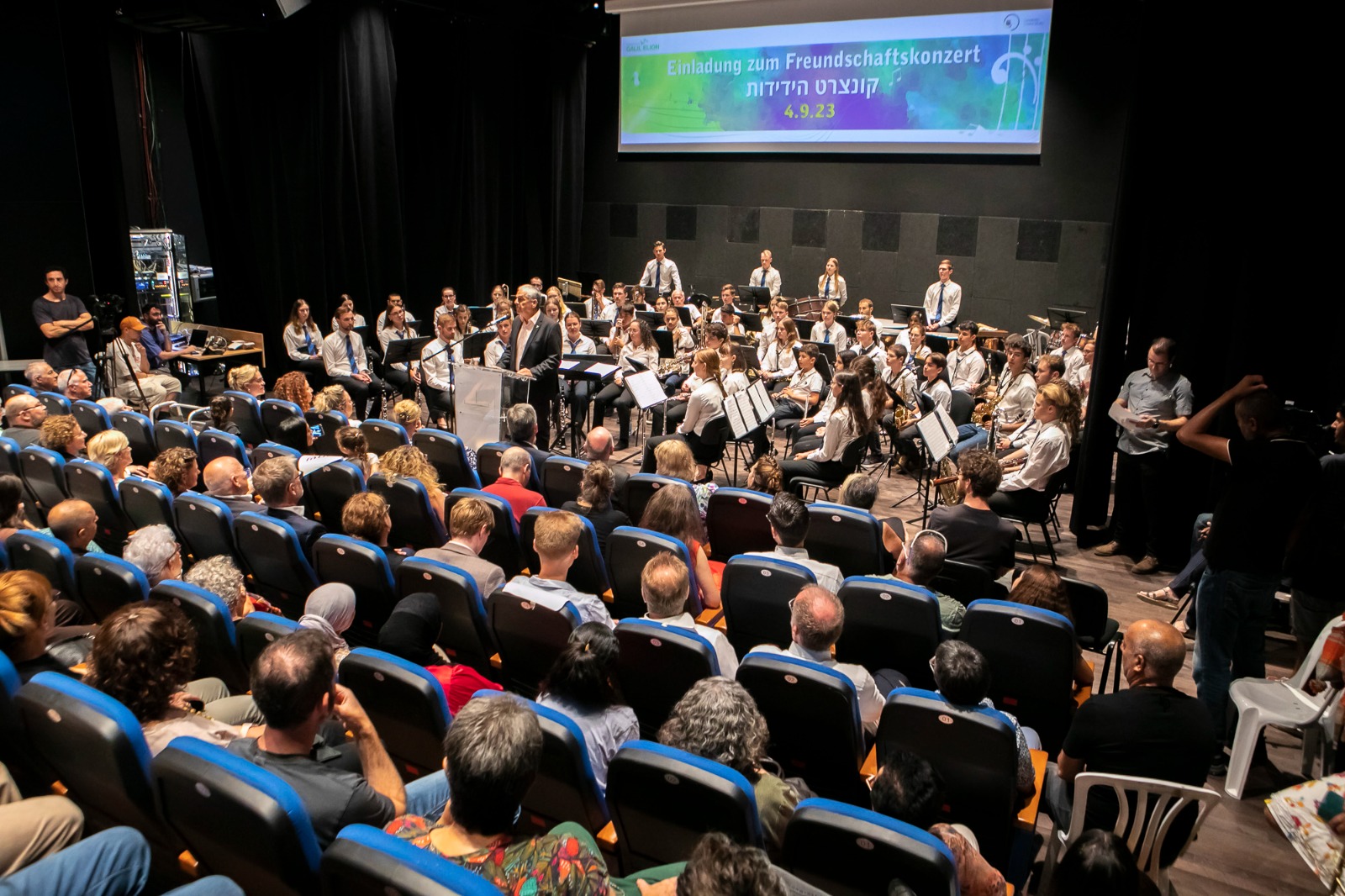 Ein Orchester spielt auf einer Bühne vor einem Publikum in einem Konzertsaal. Die Musiker sind in Weiß gekleidet, während das Publikum in bunten Kleidung sitzt und dem Konzert aufmerksam folgt. Ein Banner mit dem Titel des Freundschaftskonzerts ist im Hintergrund sichtbar.