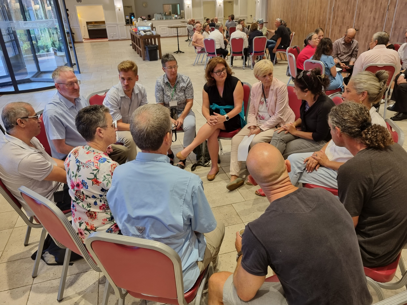 Eine Gruppe von zwölf Personen sitzt in einem Kreis auf Stühlen in einem Veranstaltungsraum. Sie diskutieren angeregt miteinander. Im Hintergrund sind andere Personen und Tische sichtbar. Der Raum hat eine freundliche, offene Atmosphäre.