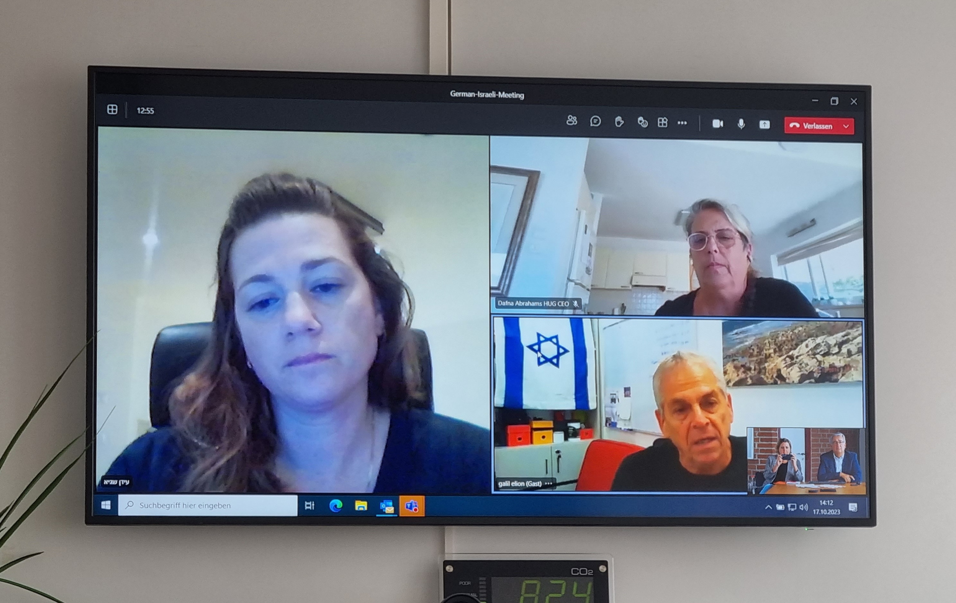 Das Bild zeigt einen virtuellen Meeting-Raum mit vier Personen. Drei von ihnen sind in separaten Fenstern angezeigt: eine Frau mit langen Haaren, eine weitere Frau mit grauen Haaren und ein Mann mit grauen Haaren. Alle blicken aufmerksam in die Kamera. Im Hintergrund sind dekorative Elemente sichtbar.