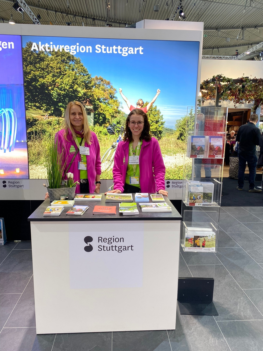 Deux femmes souriantes portent des vestes roses et se tiennent derrière un stand. Elles présentent des brochures sur la région de Stuttgart. En arrière-plan, une grande image de paysage verdoyant est visible, reflétant l'attrait touristique de la région.