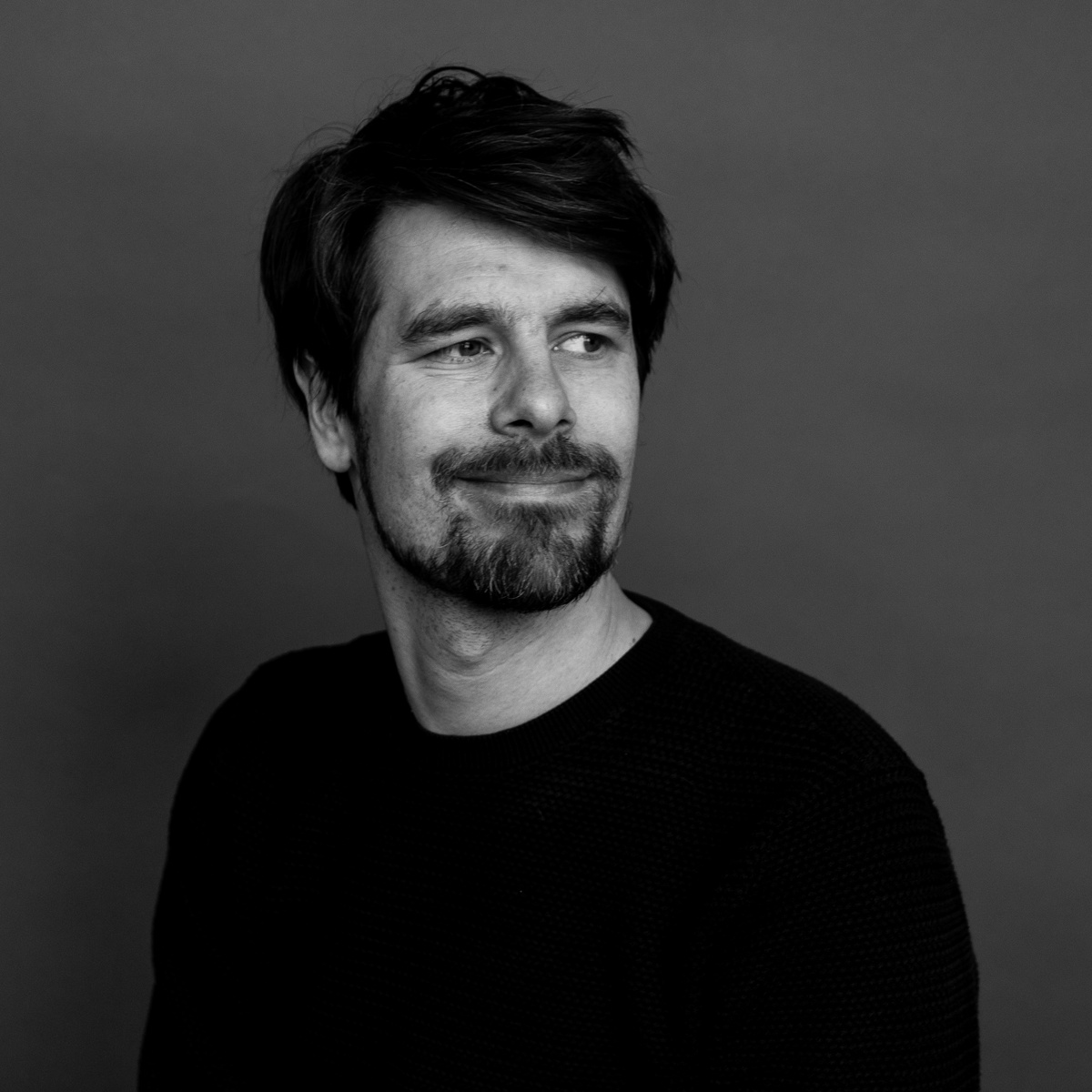 Ein Mann mit einem breiten Lächeln und einem Kurzhaarschnitt schaut leicht nach rechts. Er trägt ein schwarzes, langärmliges Oberteil und hat einen gepflegten Bart. Der Hintergrund ist einfarbig grau.