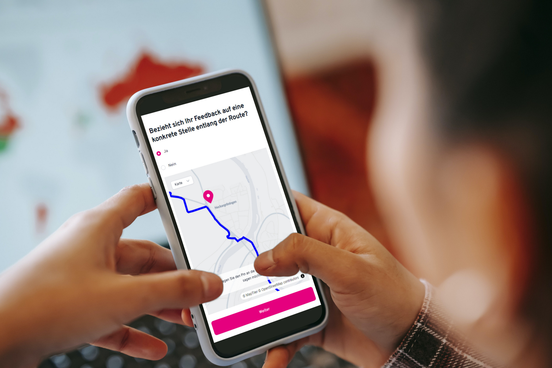 Eine Person hält ein Smartphone, auf dem eine Karte mit einer blauen Route und einem pinken Standortmarker angezeigt wird. Der Bildschirm zeigt eine Anfrage zur Rückmeldung zu einer möglichen Route an. Im Hintergrund ist ein Laptop mit weiteren Karten sichtbar.