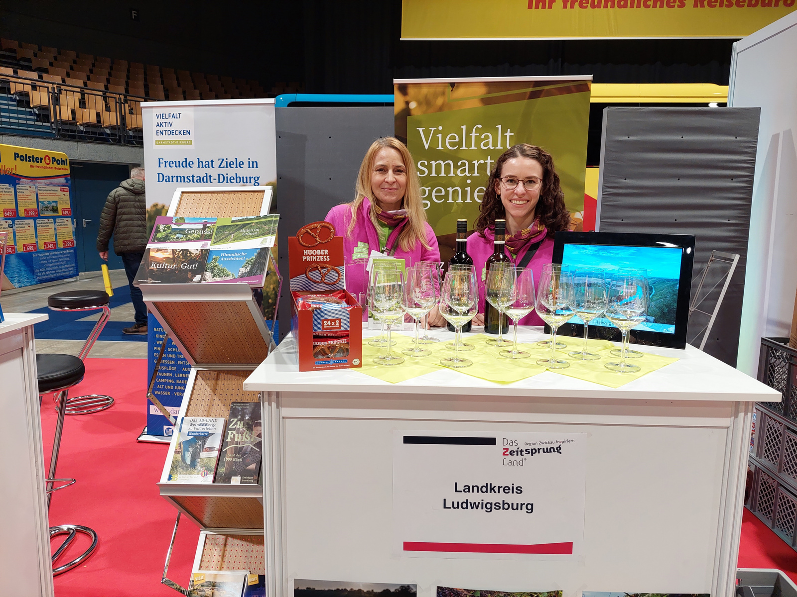 Deux femmes se tiennent derrière un stand d'exposition avec des verres à vin au premier plan. Le stand représente le Landkreis Ludwigsburg, et des brochures sont visibles sur la table, affichant des informations sur la région. Une ambiance accueillante et informative se dégage de l'espace d'exposition.