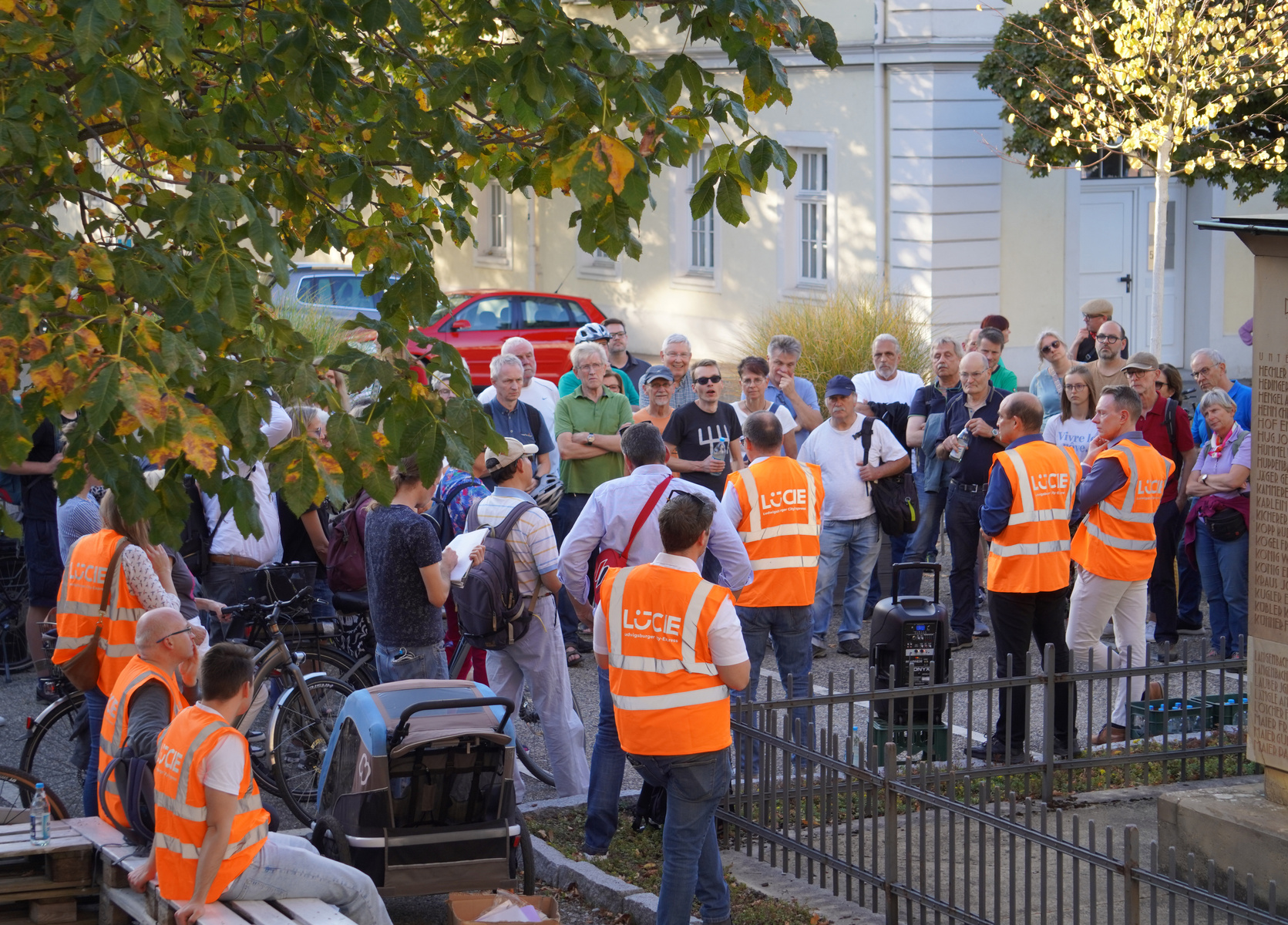 A group of people is gathered outdoors, some wearing bright orange vests labeled "LUCE." The setting appears to be an event or community meeting, with a mixture of participants engaging in conversation amidst trees and buildings in the background.