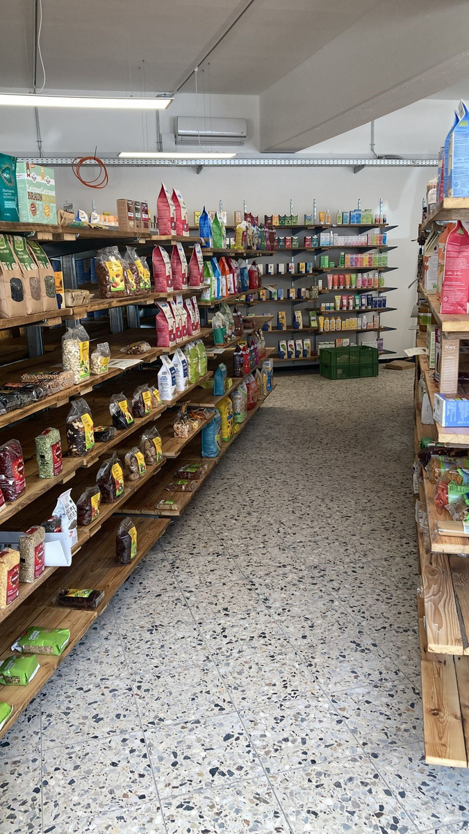 Ein Innenansicht eines Lebensmittelladens mit Holzregalen auf beiden Seiten. Die Regale sind mit verschiedenen Verpackungen gefüllt, darunter Snacks, Trockenfrüchte und andere Produkte. Der Abstand in der Mitte bietet Platz für das Flanieren zwischen den Regalen.