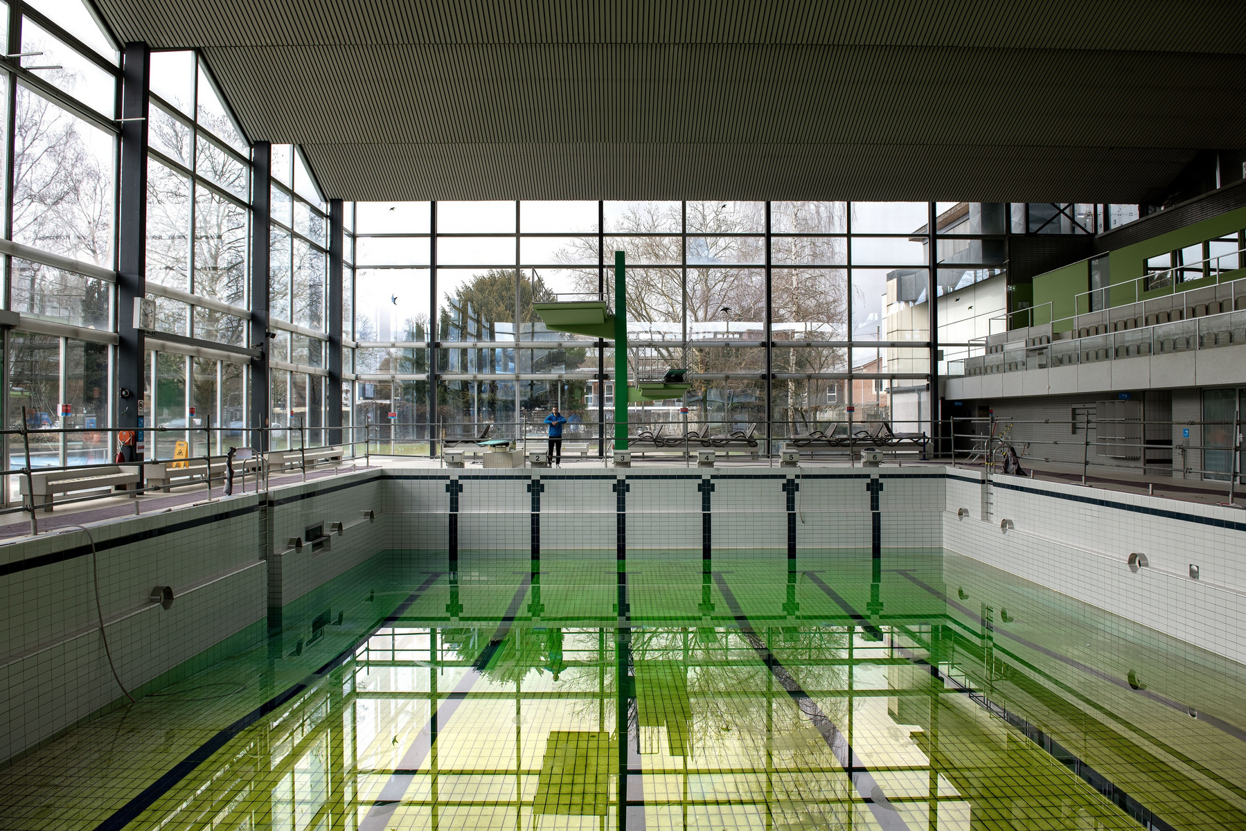 Das Bild zeigt ein leeres Schwimmbad mit grünem Wasser und einem Sprungturm. Große Fenster lassen Tageslicht herein, und im Hintergrund ist eine Person zu sehen, die sich in der Schwimmhalle aufhält. Die Umgebung wirkt modern und geräumig.
