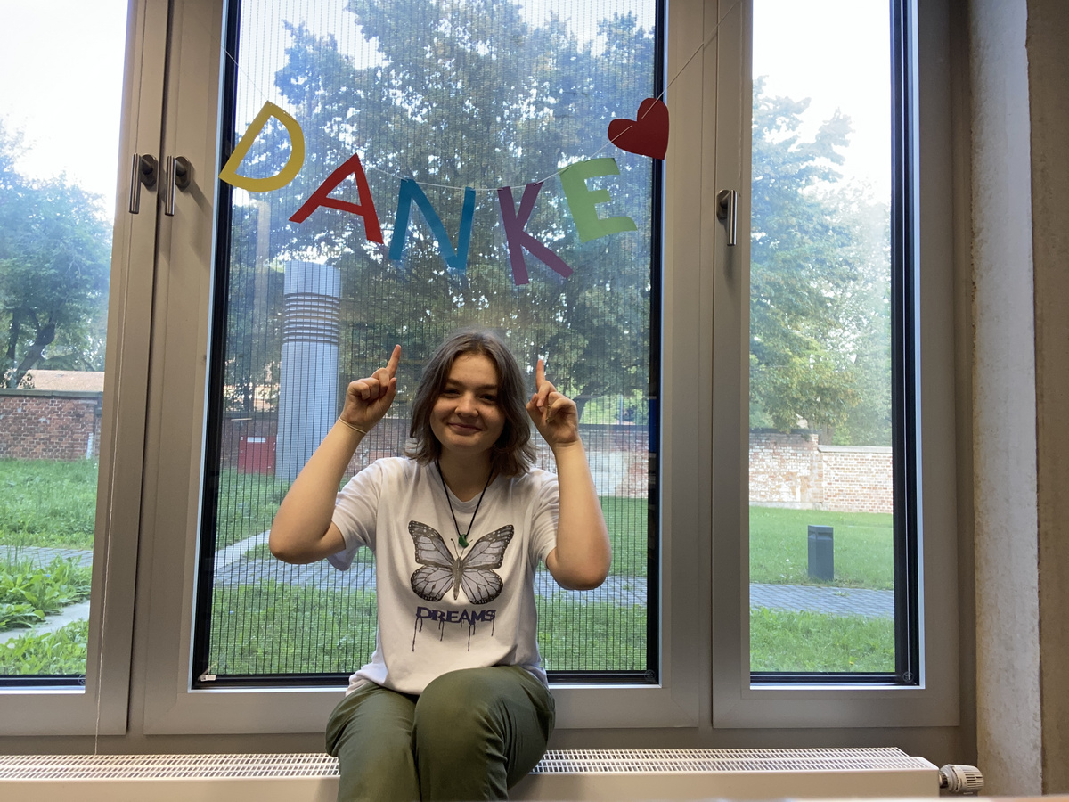 Eine lächelnde Person zeigt mit beiden Händen nach unten auf das Wort "DANKE", das farbenfroh am Fenster geschrieben ist. Im Hintergrund sind Bäume und Rasen sichtbar.