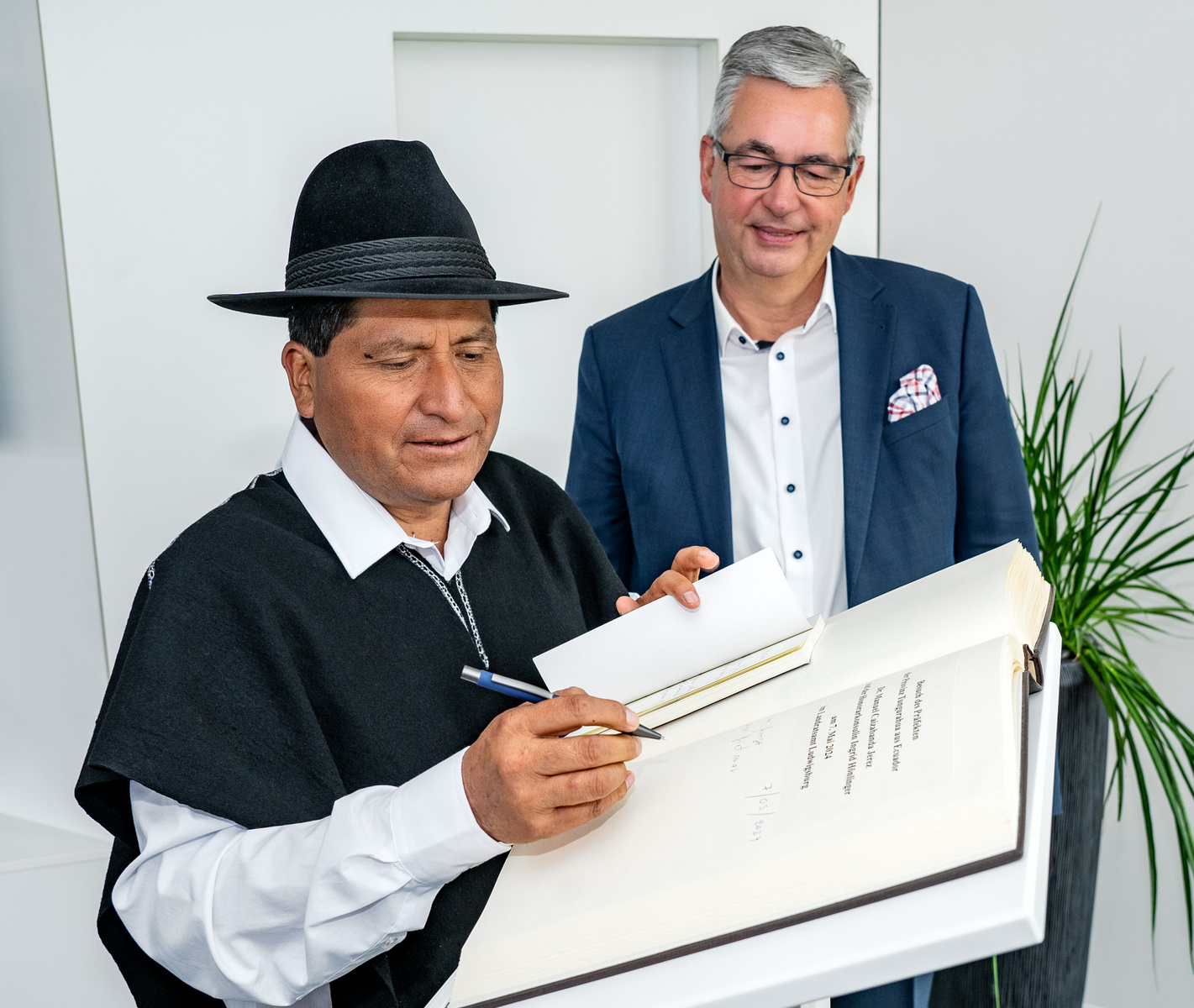 Due uomini sono presenti in un ambiente moderno. Uno, vestito con un poncho e un cappello, firma un documento con una penna. L'altro, in abito elegante, osserva. Una pianta decorativa è visibile sullo sfondo.