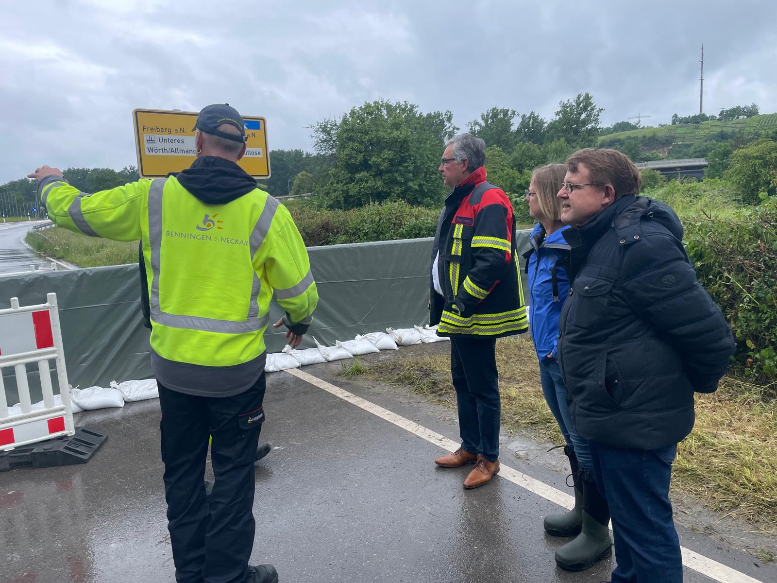 Vier Personen stehen an einer abgesperrten Straße und sprechen miteinander. Eine Person in Warnjacke zeigt auf ein Schild. Die Umgebung ist bewaldet, der Himmel ist bewölkt und es scheint, als ob es regnet.