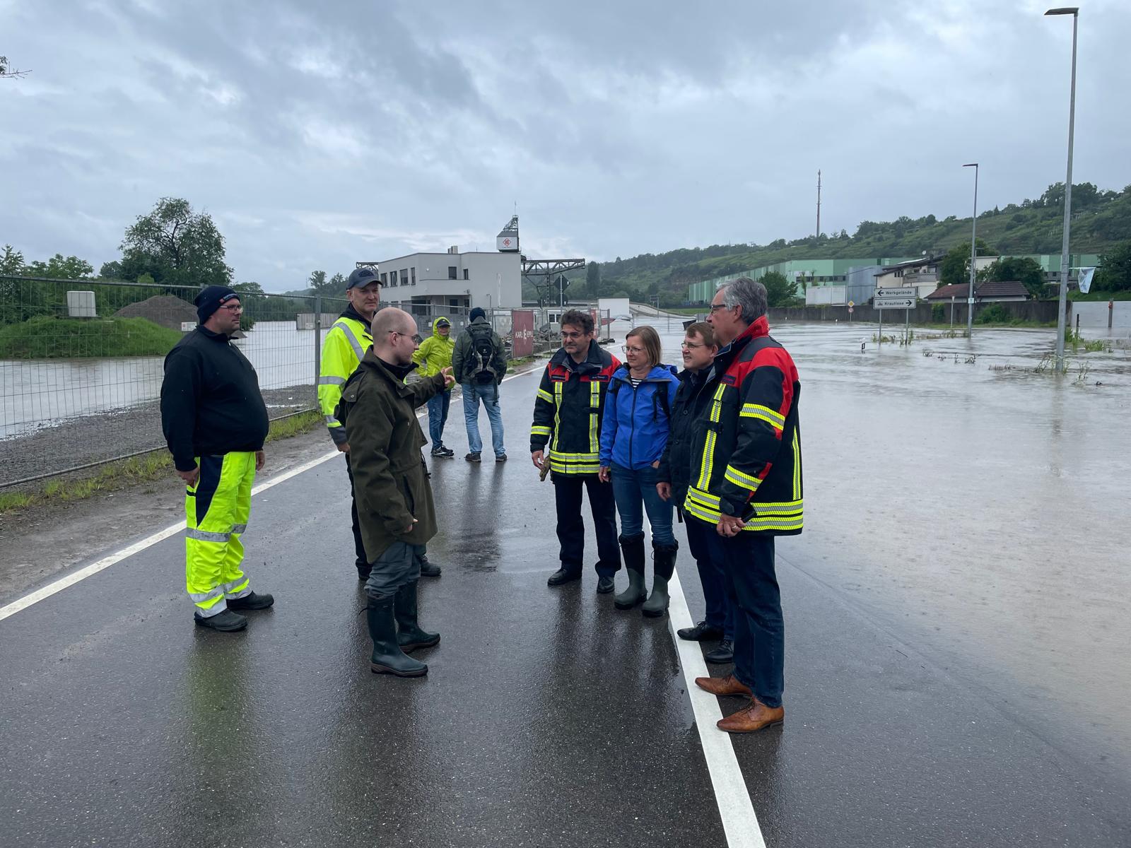 Eine Gruppe von Einsatzkräften und Behördenvertretern steht an einer überschwemmten Straße. Sie diskutieren die Situation während regnerischen Wetterbedingungen. Im Hintergrund sind Wasseransammlungen und einige Gebäude sichtbar. Personen tragen wetterfeste Kleidung und Sicherheitsausrüstung.