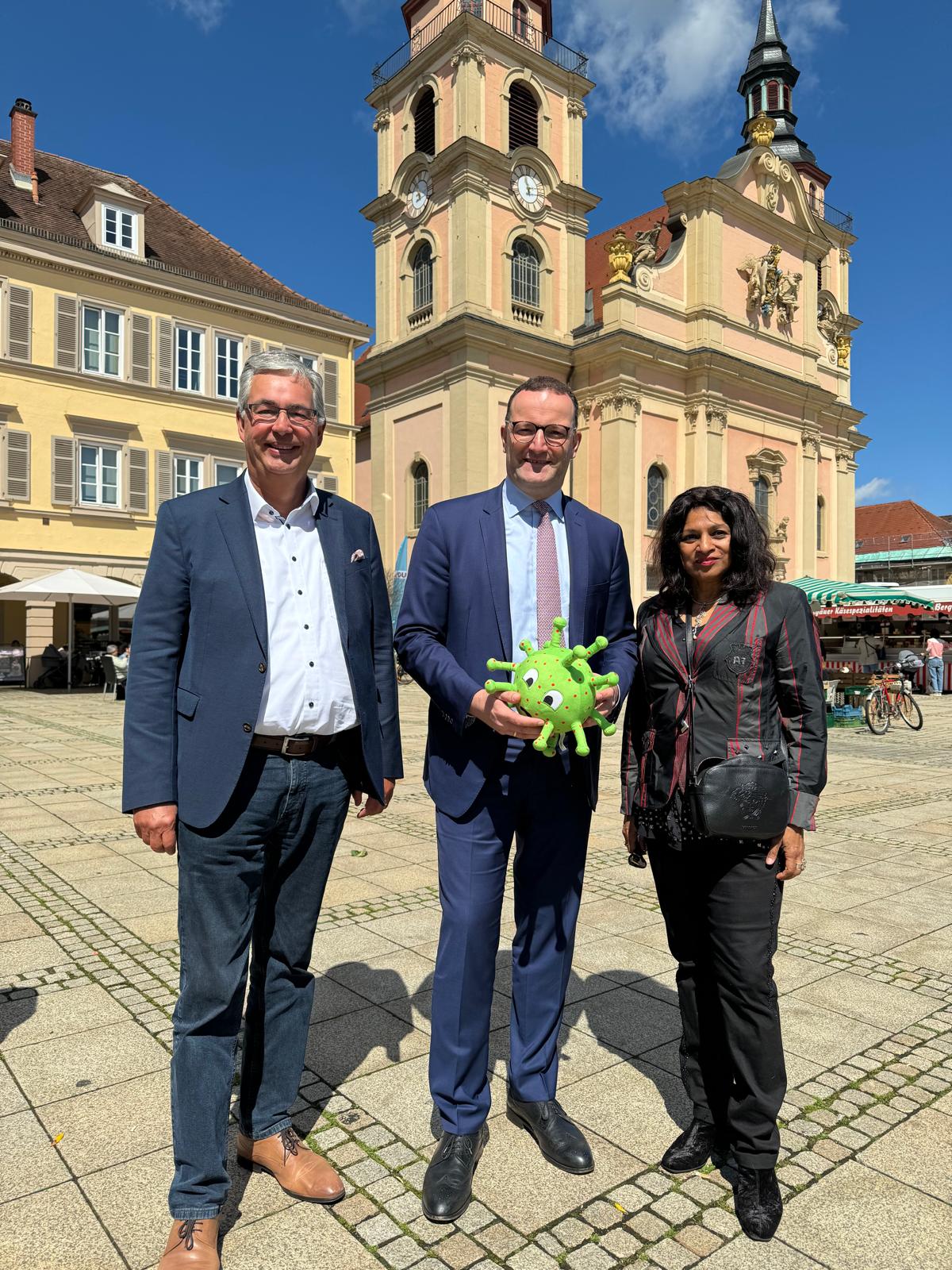Drei Personen stehen auf einem Platz vor einem historischen Gebäude. Der Mann in der Mitte hält ein grünes, plüschiges Virusmodell. Die andere zwei Personen stehen zu seiner linken und rechten Seite und blicken zur Kamera. Helles Wetter mit blauen Himmel.