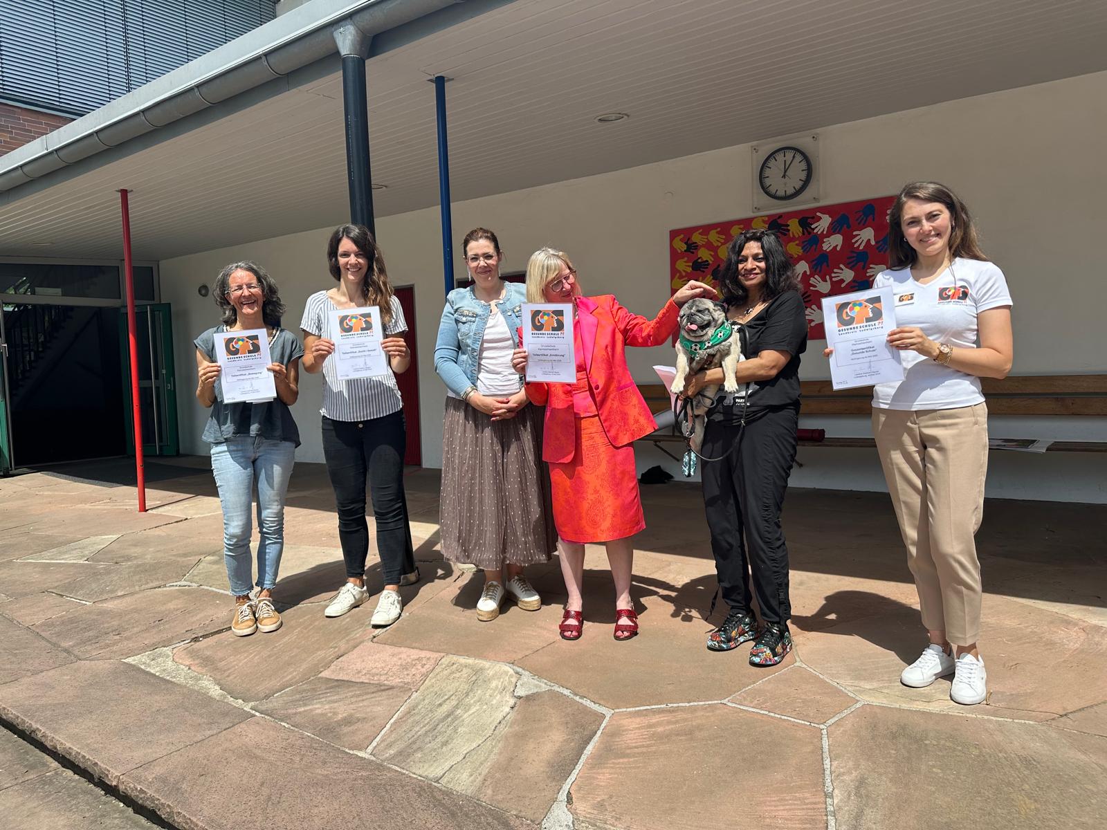 Sechs Personen stehen in einer schattigen Umgebung und halten jeweils ein Dokument in der Hand. Eine Frau in einem roten Outfit hält einen Hund. Im Hintergrund sind Bänke und bunte Kunstwerke zu sehen.