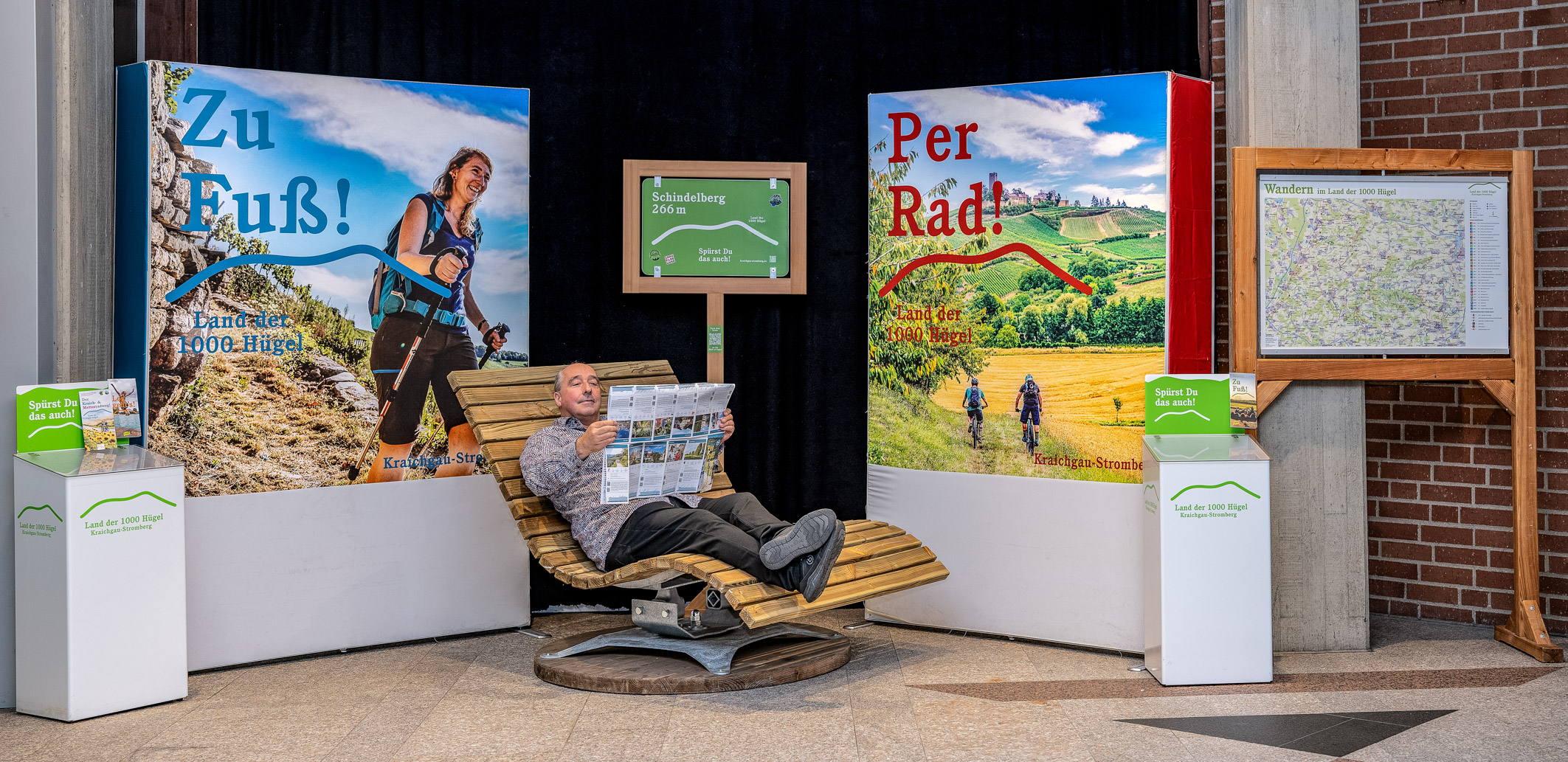 Eine Ausstellung mit dem Thema Outdoor-Aktivitäten zeigt zwei großflächige Plakate: eines mit der Aufschrift „Zu Fuß!“ und das andere „Per Rad!“. In der Mitte sitzt ein Mann auf einer Liege und betrachtet eine Reisebroschüre. Im Hintergrund sind Angebote für Wander- und Radfahrtouren dargestellt.