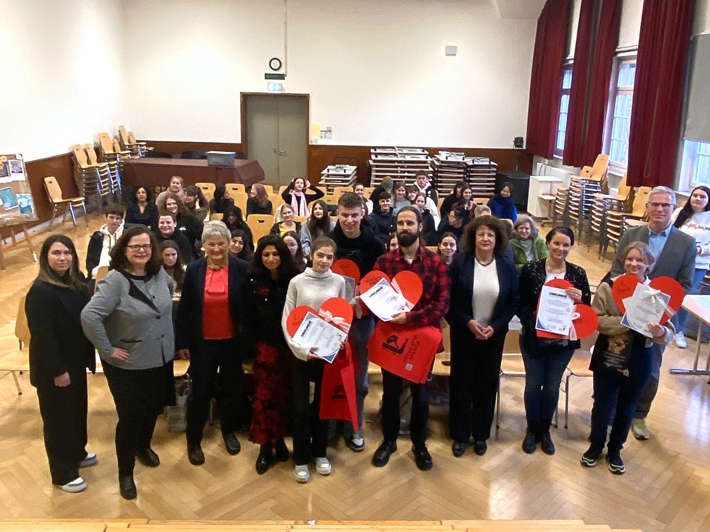 Eine Gruppe von Menschen steht in einem Raum, umgeben von Stühlen und Tischen. Viele halten rote Herzen und Zertifikate in der Hand. Die Anwesenden lächeln, während sie für ein Gruppenfoto posieren. Der Raum hat große Fenster und eine freundliche Atmosphäre.