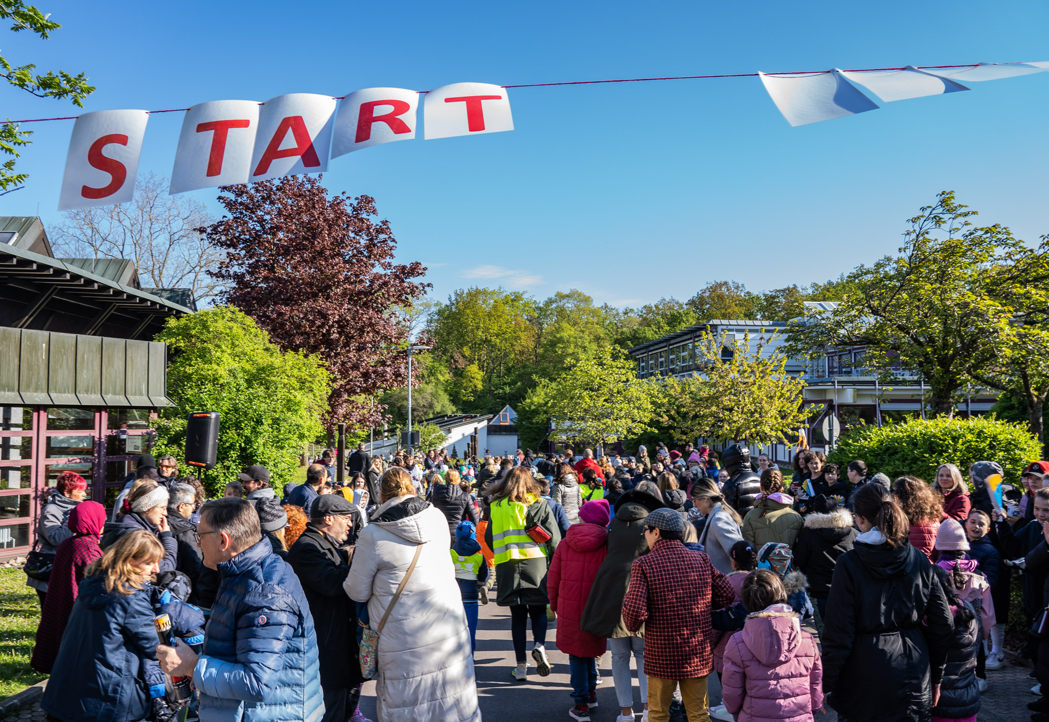 Mehrere Menschen versammeln sich an einem sonnigen Tag unter einem großen "START"-Schild. Die Umgebung ist grün, mit Bäumen und Gebäuden im Hintergrund. Die Teilnehmer wirken bereit, an einer Veranstaltung oder einem Wettlauf teilzunehmen.
