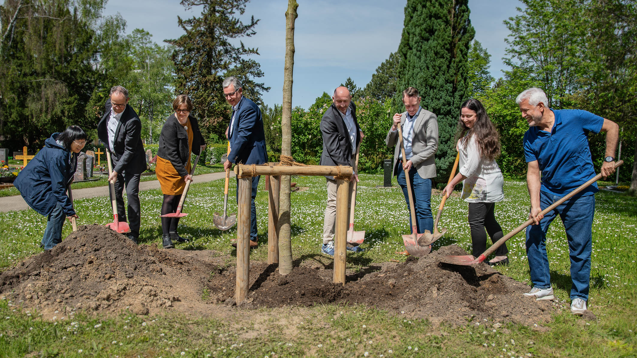 A group of eight people gathers outdoors, using shovels to plant a young tree. They are standing on soil mounds in a grassy area surrounded by trees. The scene captures a collaborative effort for environmental stewardship.