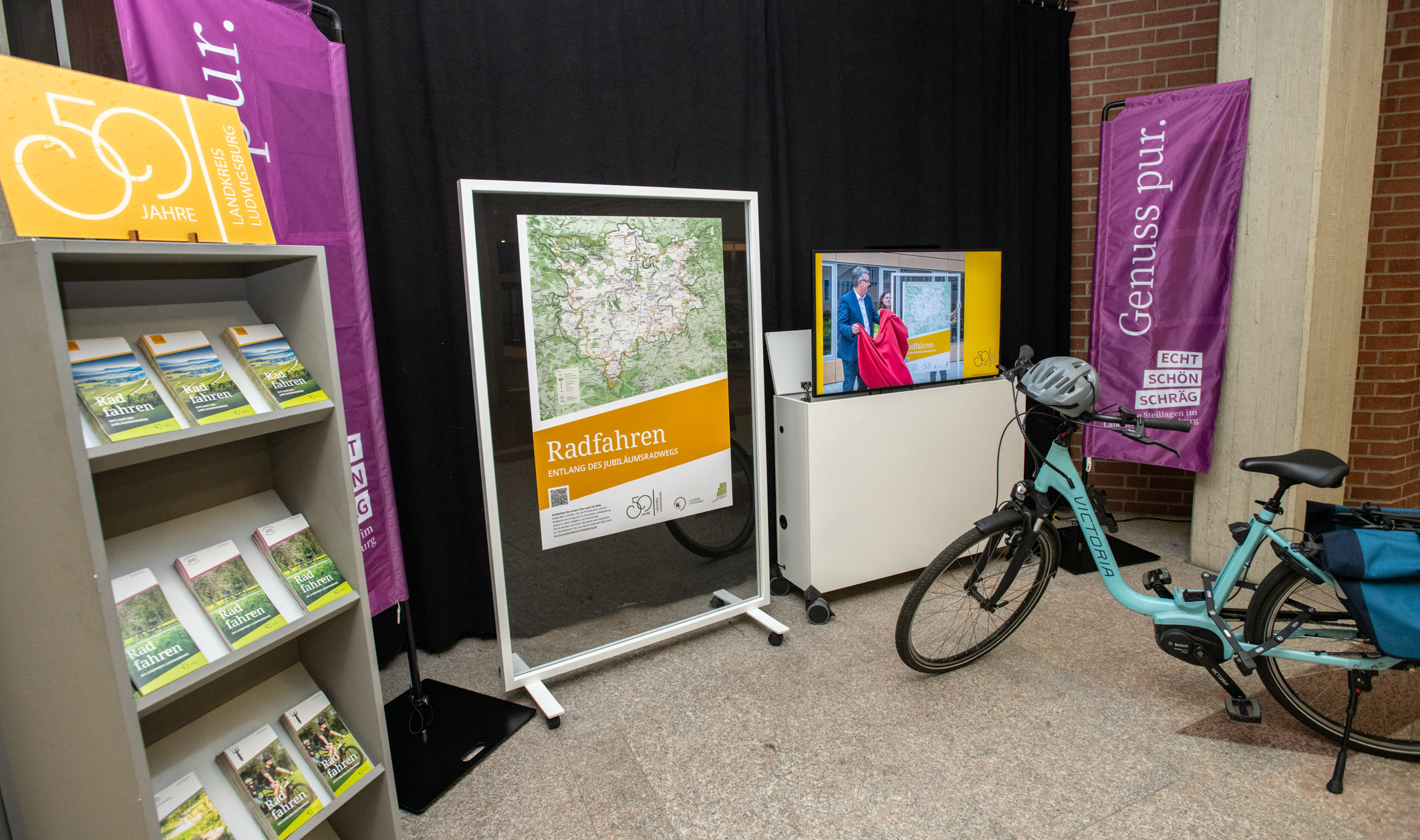 Eine Ausstellung über Radfahren mit einem großen Infobreich, der eine Karte zeigt. Neben dem Infobereich stehen Fahrräder und ein Präsentationsbildschirm. In der Nähe befinden sich Informationsbroschüren zum Thema Radfahren. Im Hintergrund sind dekorative Displays zu sehen.