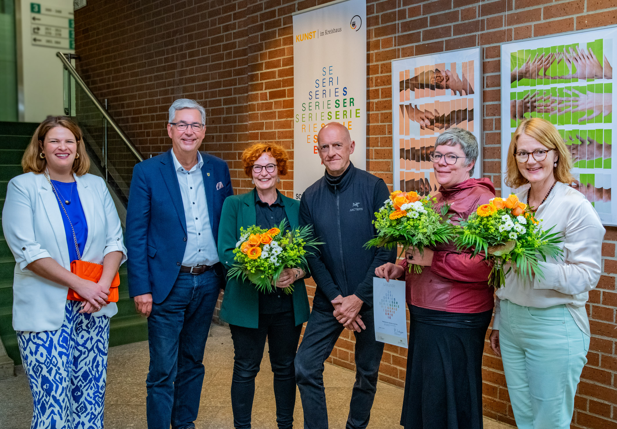 Eine Gruppe von sechs Personen steht vor einer Wand mit Kunstwerken. Alle halten Blumensträuße. Zwei Personen tragen formelle Kleidung, während die anderen lässiger gekleidet sind. Im Hintergrund ist eine Informationswand zur Ausstellung zu sehen. Die Stimmung ist festlich und freundlich.