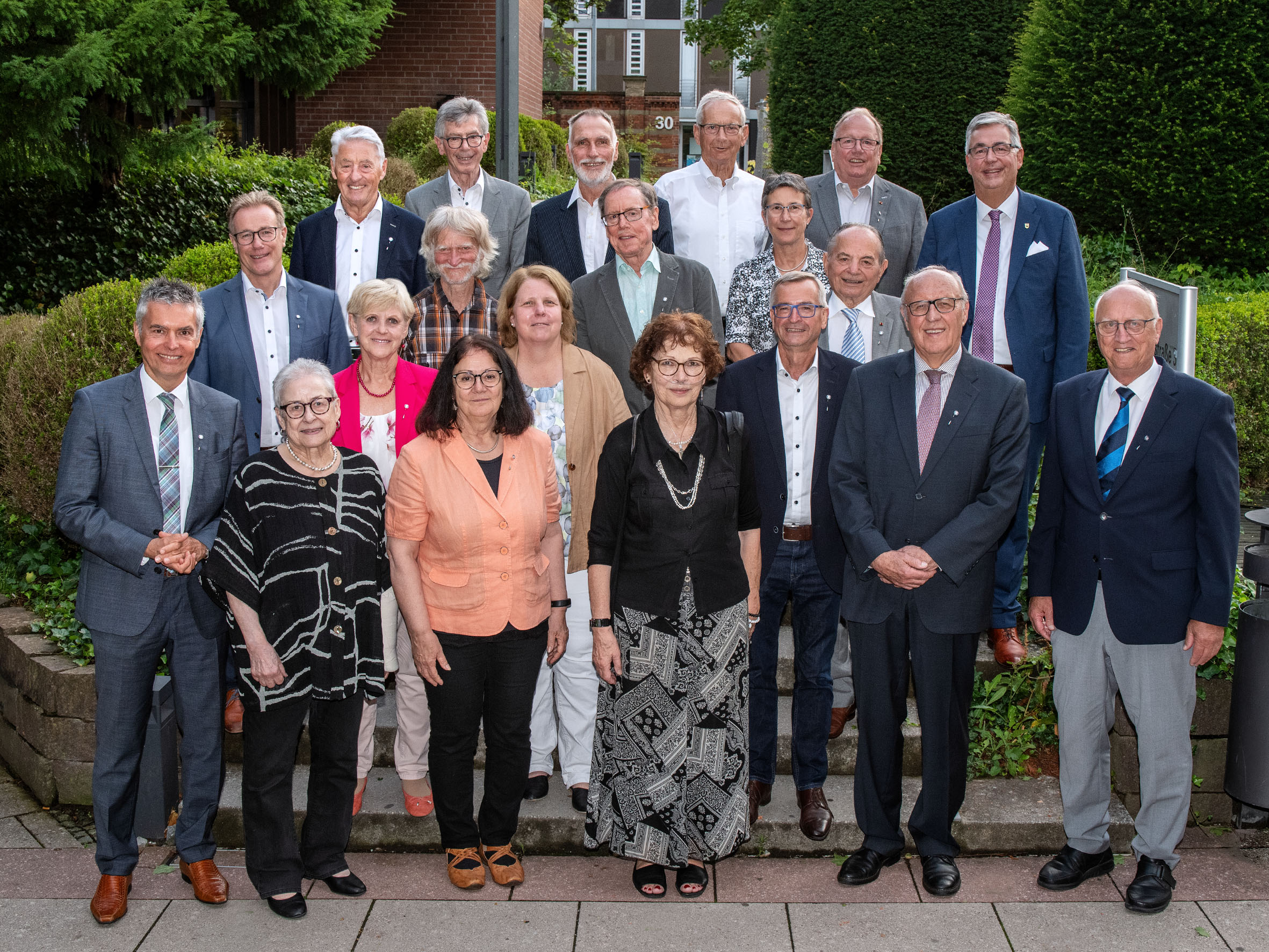 Eine Gruppe von 20 Personen steht im Freien in einer Gartenumgebung. Die Menschen sind in formeller Kleidung gekleidet, und einige lächeln in Richtung der Kamera. Im Hintergrund sind Büsche und ein Gebäude zu sehen.