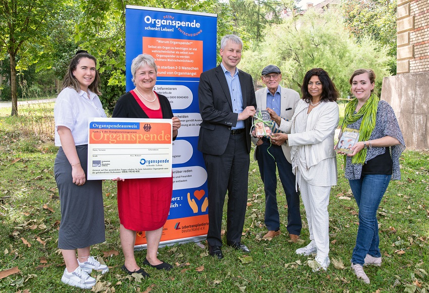 Eine Gruppe von sechs Personen steht im Freien und hält einen großen Scheck zur Organspende. Im Hintergrund ist ein Banner mit Informationen zur Organspende sichtbar. Alle Personen wirken freundlich und engagiert in ihrer Unterstützung für die Organspendebewegung.
